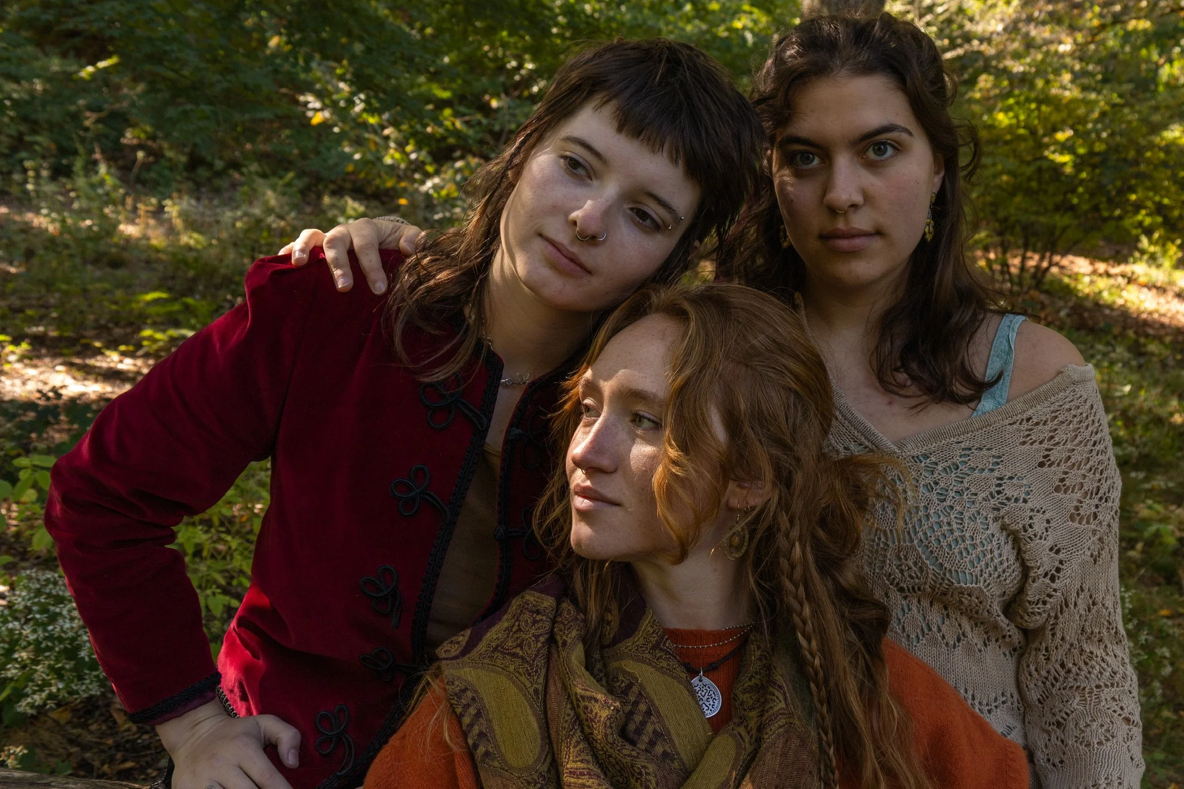 Three women with diverse appearances posed closely together outdoors in a wooded area, with sunlight filtering through the trees.