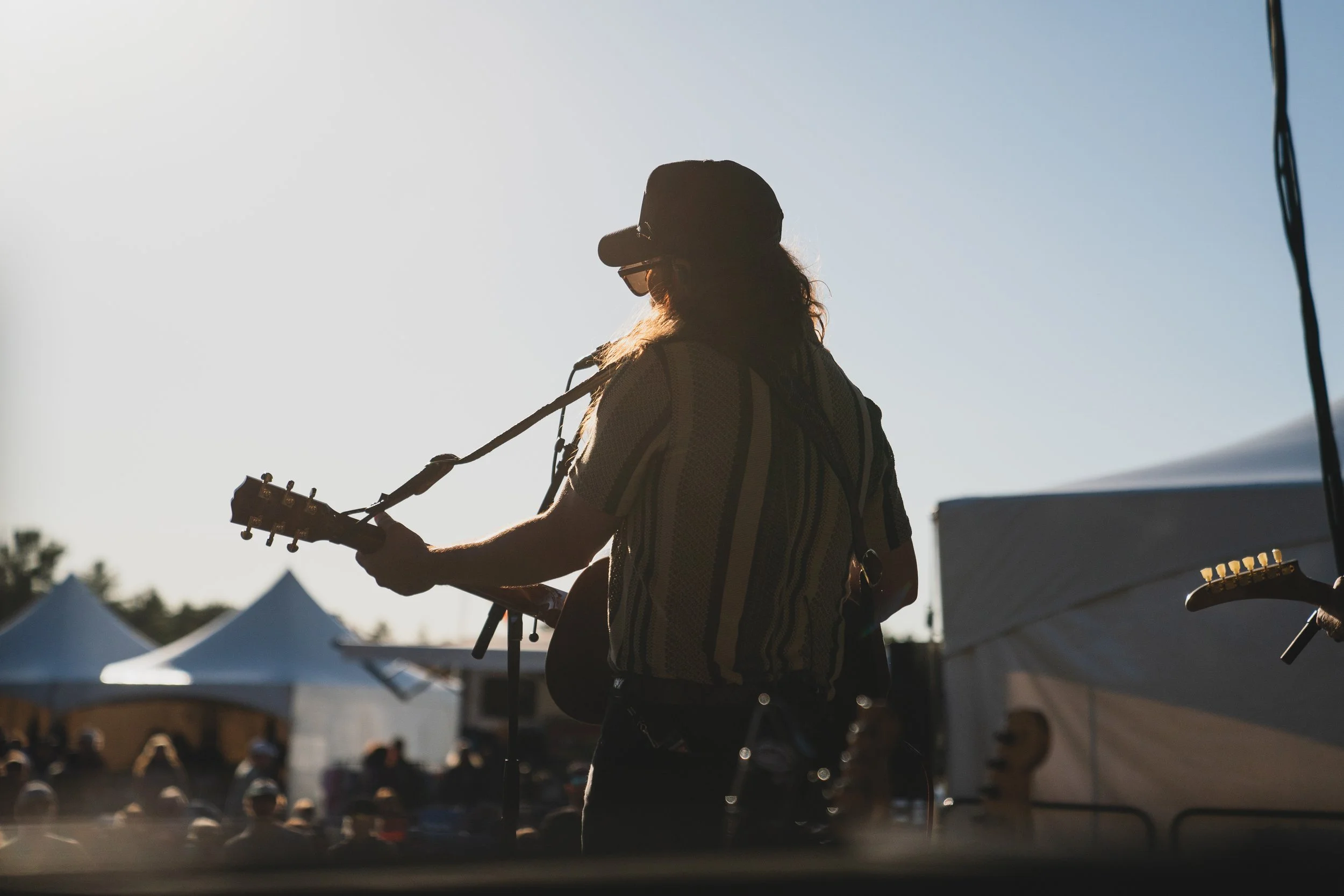 Silhouette of a person playing an acoustic guitar outdoors during late afternoon or evening, with tents and a crowd visible in the background.