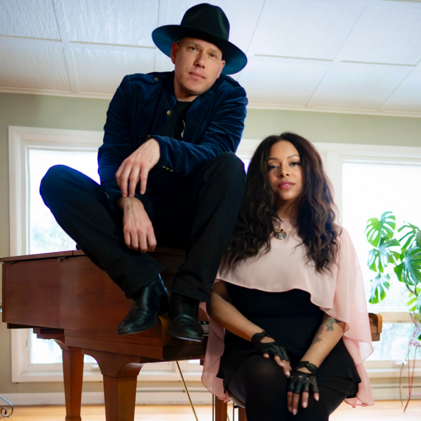 A man wearing a black wide-brimmed hat and dark clothing sits on a piano, and a woman with wavy dark hair, wearing a light pink cape, black dress, and gloves, sits beside him in a room with large windows and a green plant.