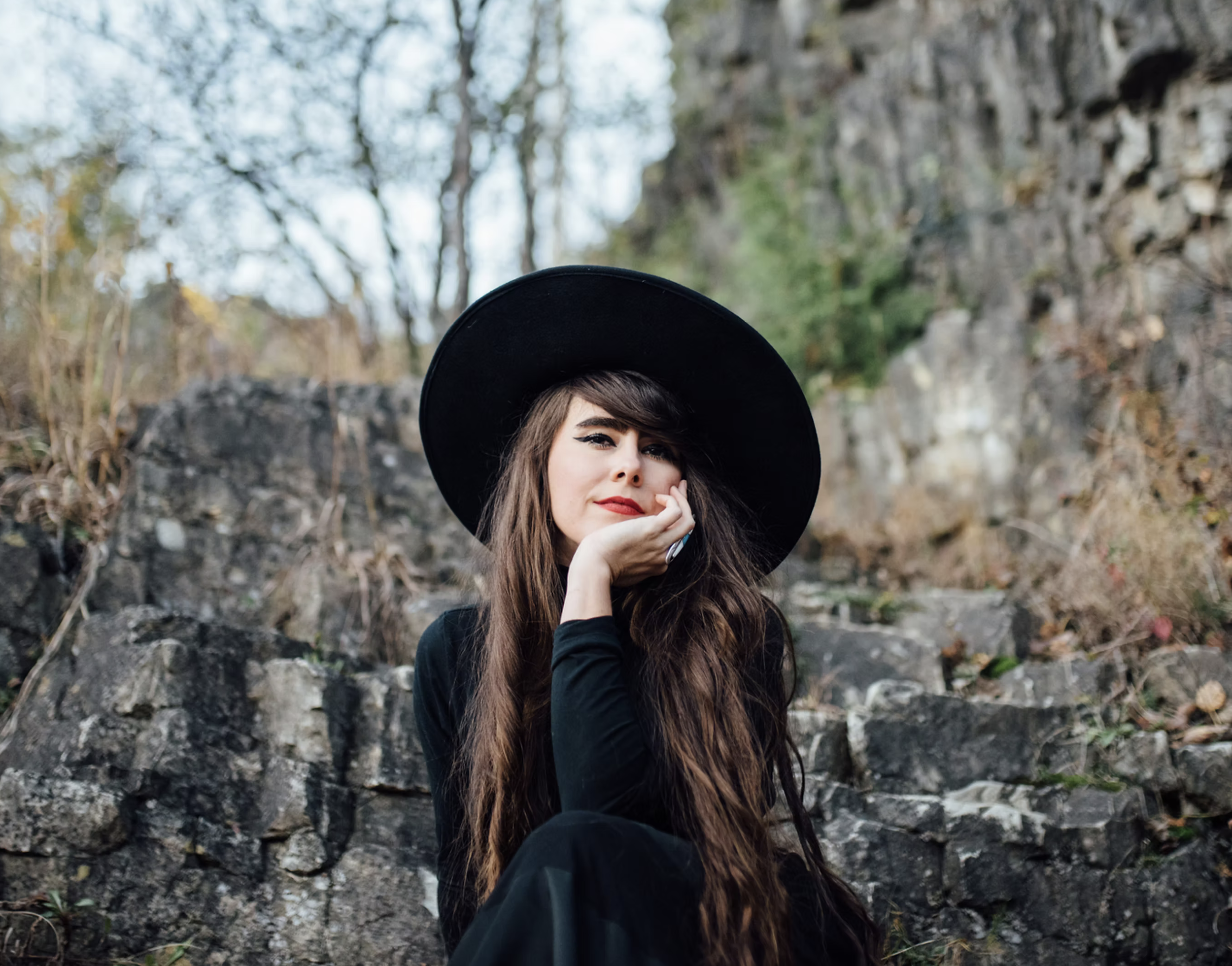 A woman with long brown hair, red lipstick, wearing a black wide-brimmed hat and black clothing, sitting outdoors on rocks with a rocky cliff and trees in the background.