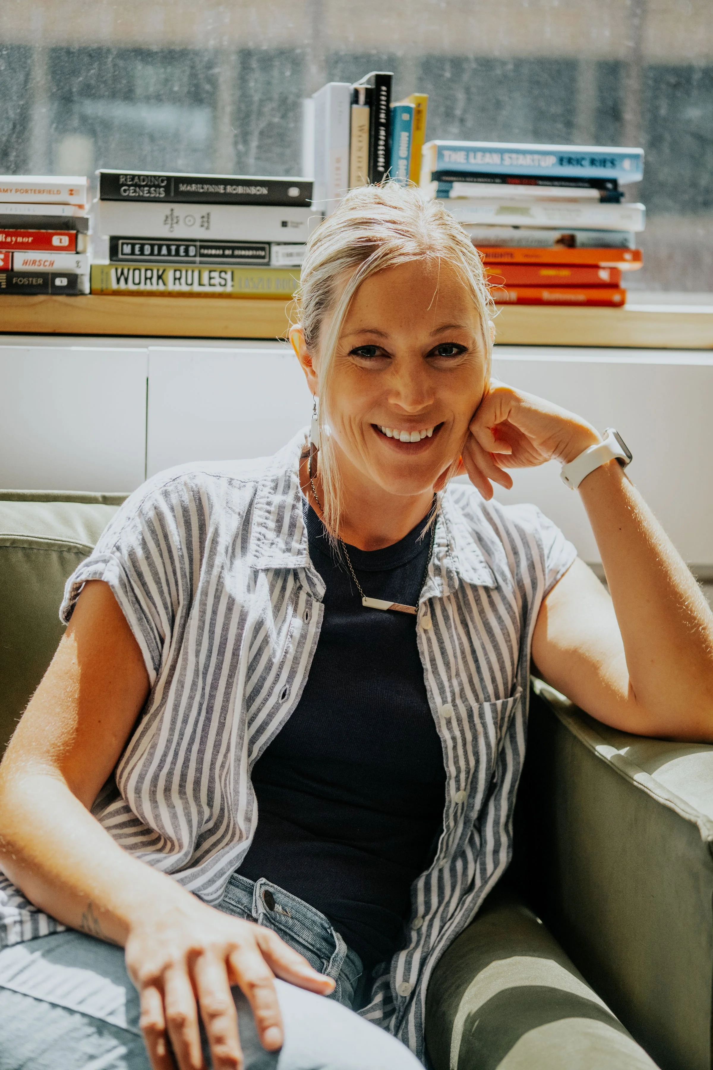 A woman sitting on a green couch, smiling, with her elbow resting on the armrest and her chin on her hand, in front of a window with books on a shelf behind her.