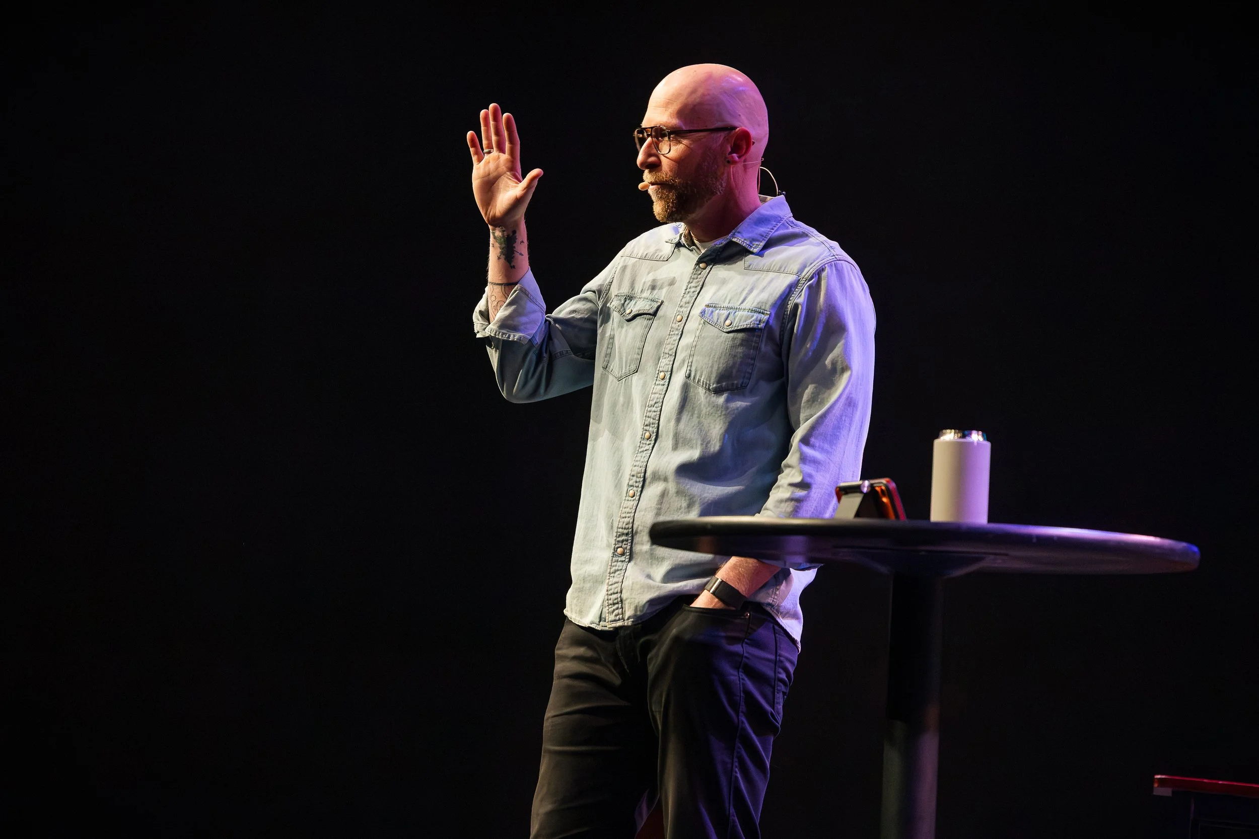A man giving a presentation to an audience seated at tables in a conference room. A large screen behind him displays a book titled 'Without' and a coffee mug. Attendees are listening attentively.