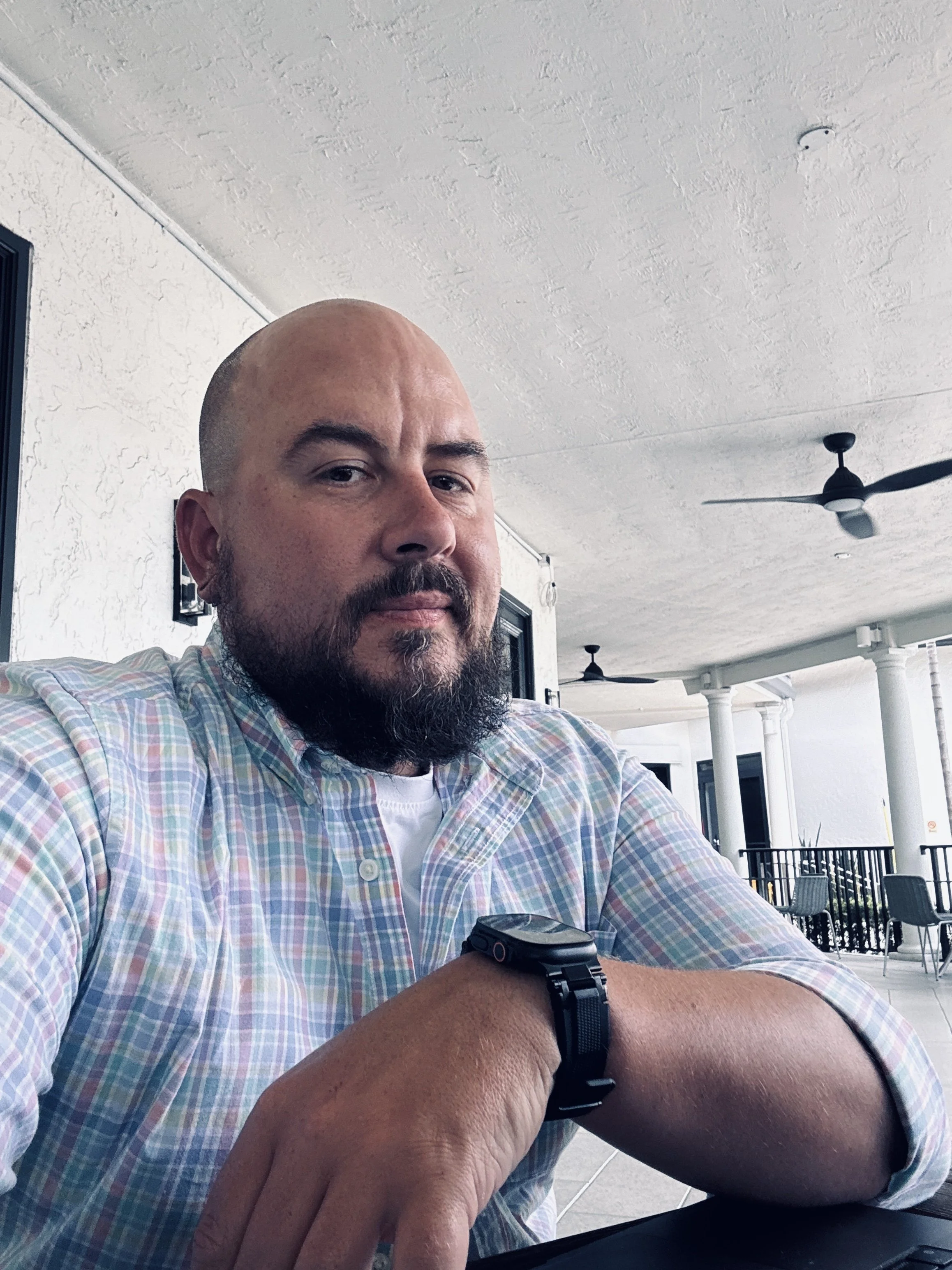 A man with a bald head and beard, wearing a checkered shirt and a watch, sitting outdoors on a patio with ceiling fans and patio furniture.