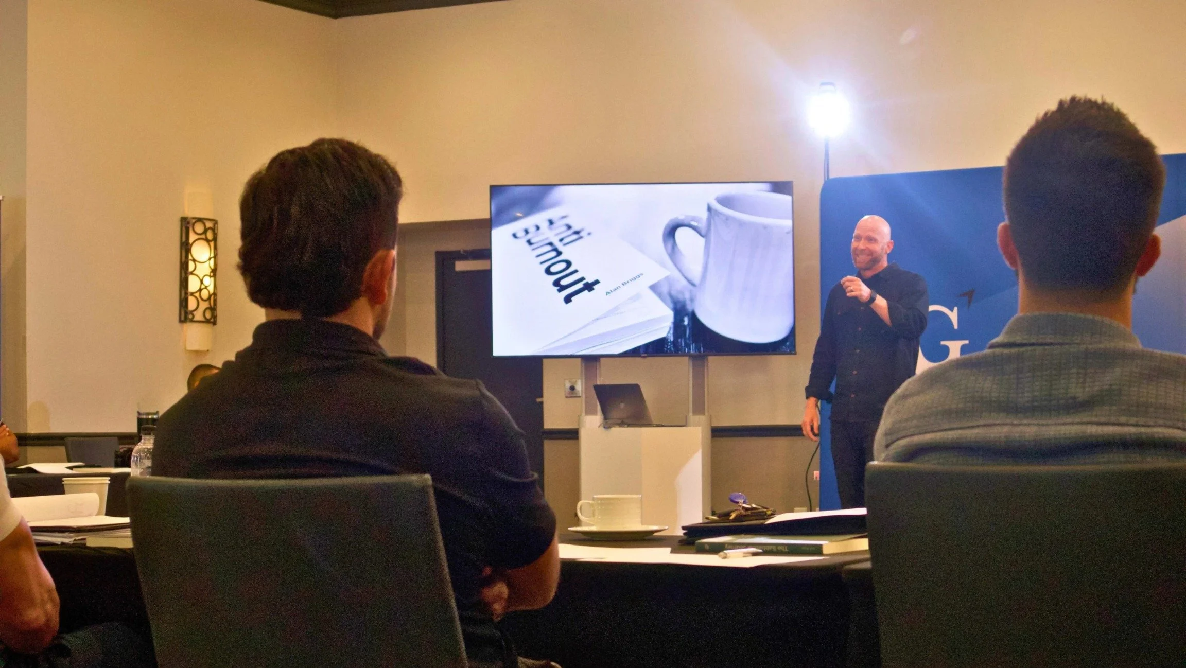 A man giving a presentation to an audience seated at tables in a conference room. A large screen behind him displays a book titled 'Without' and a coffee mug. Attendees are listening attentively.