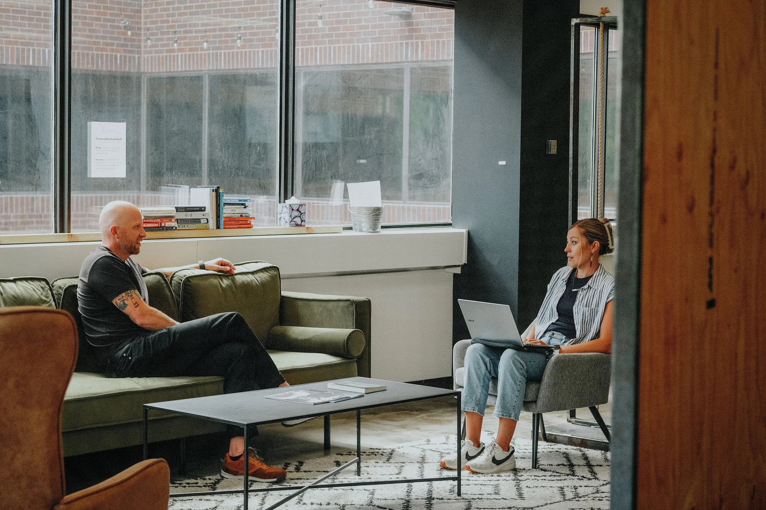 A man and a woman are sitting in a modern office lounge, having a conversation. The man is on a green couch, casually dressed, and the woman sits on a gray chair with a laptop on her lap. There is a black coffee table with magazines and a patterned rug on the floor. Large windows and a windowsill with books and decorative items are visible in the background.