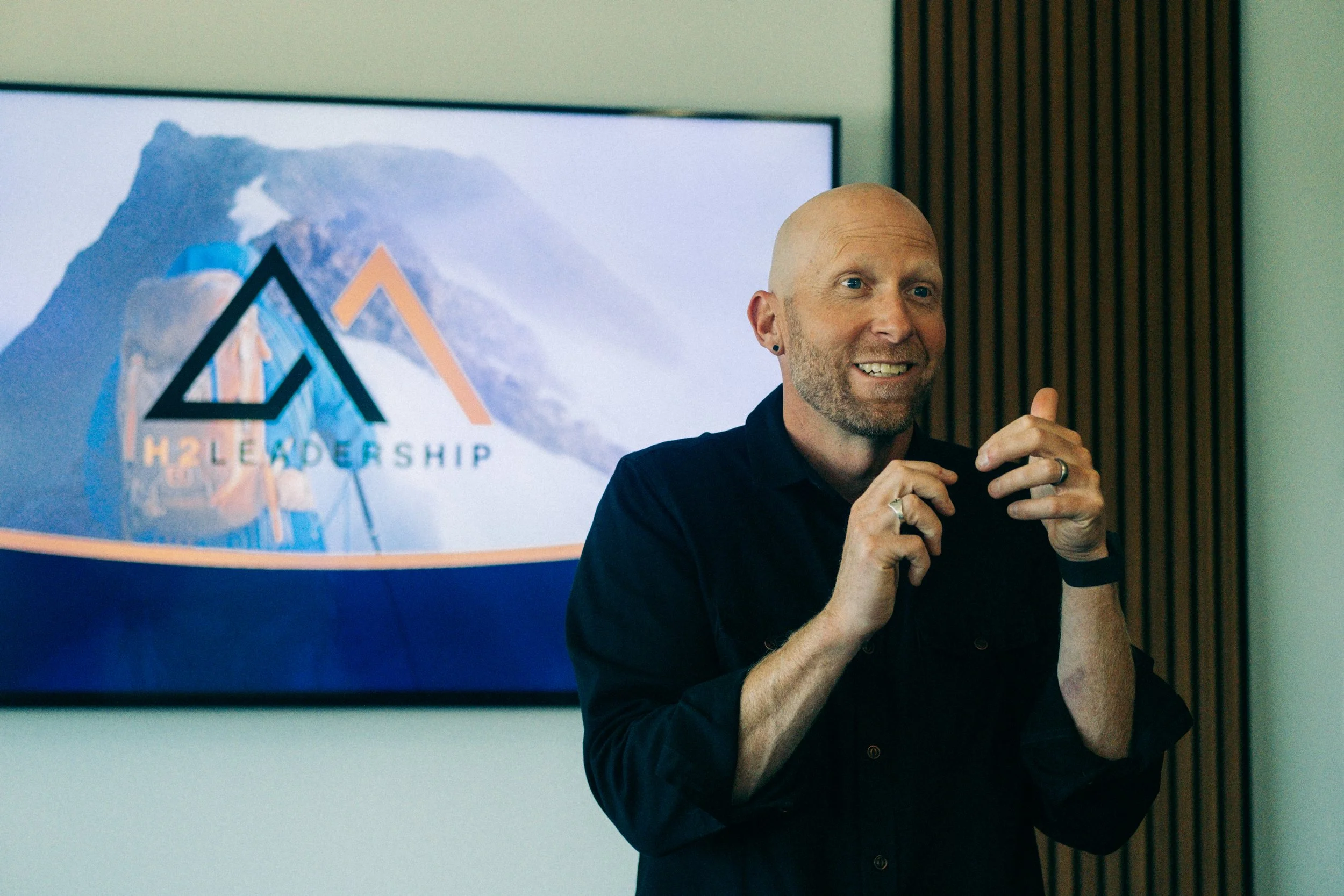 Man presenting in front of a screen with logos of H2 Leadership, smiling, with a wooden wall panel to the right.