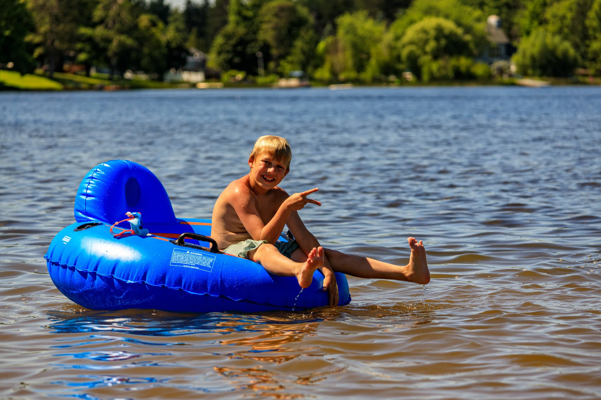 A young boy with blond hair is sitting in a blue inflatable boat on a lake, making a peace sign with his hand and smiling at the camera.
