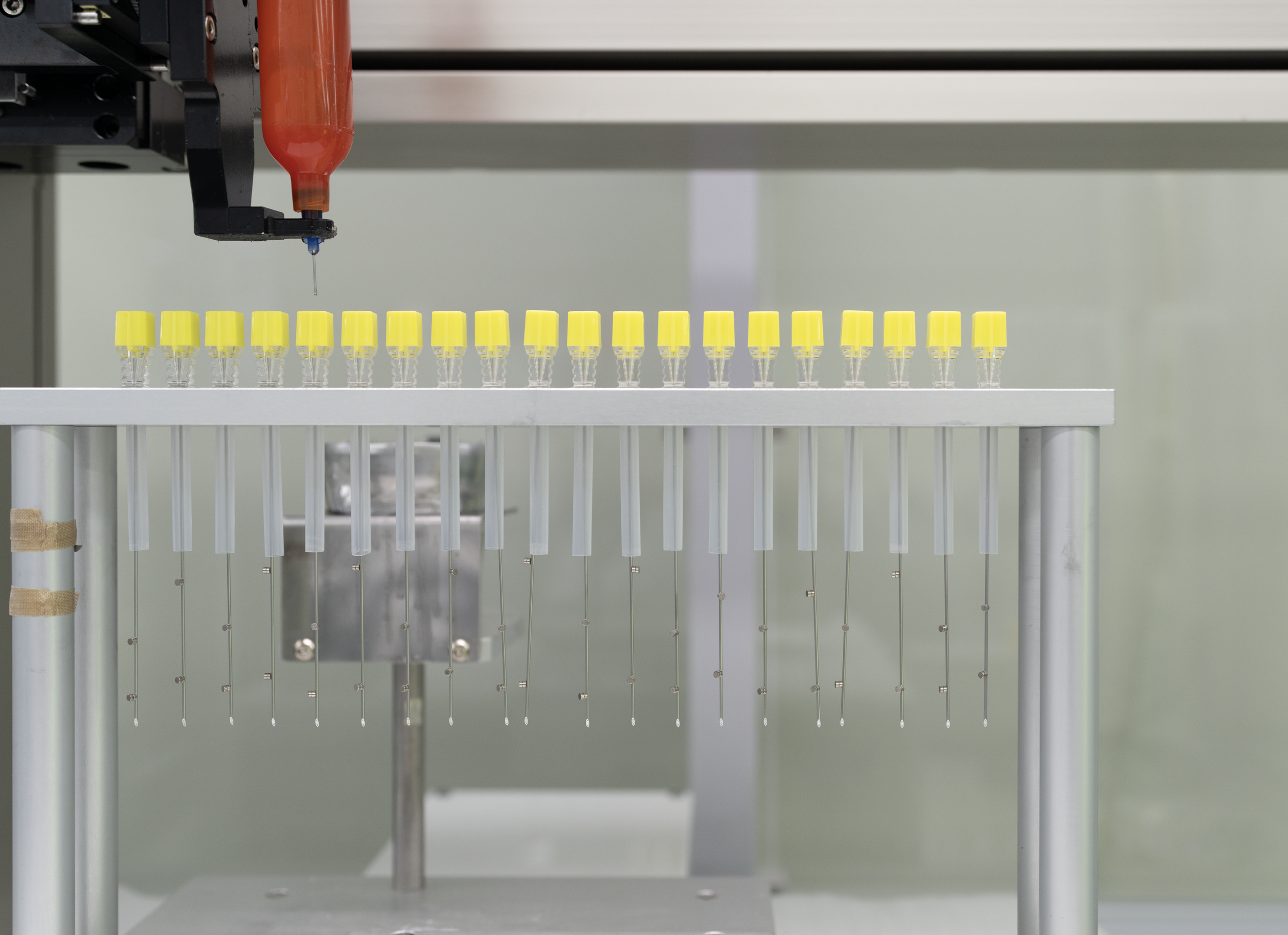 Close-up of laboratory equipment with multiple sample vials and a pipette dispenser over a rack of small yellow-topped test tubes.