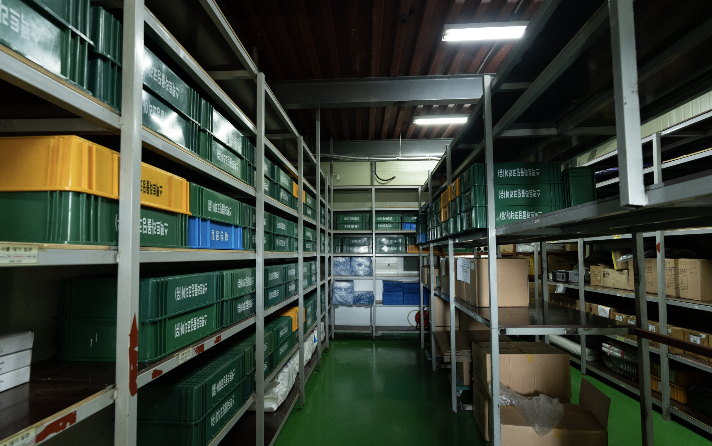 Organized storage room with metal shelving units filled with color-coded plastic bins and cardboard boxes in a clean industrial facility.