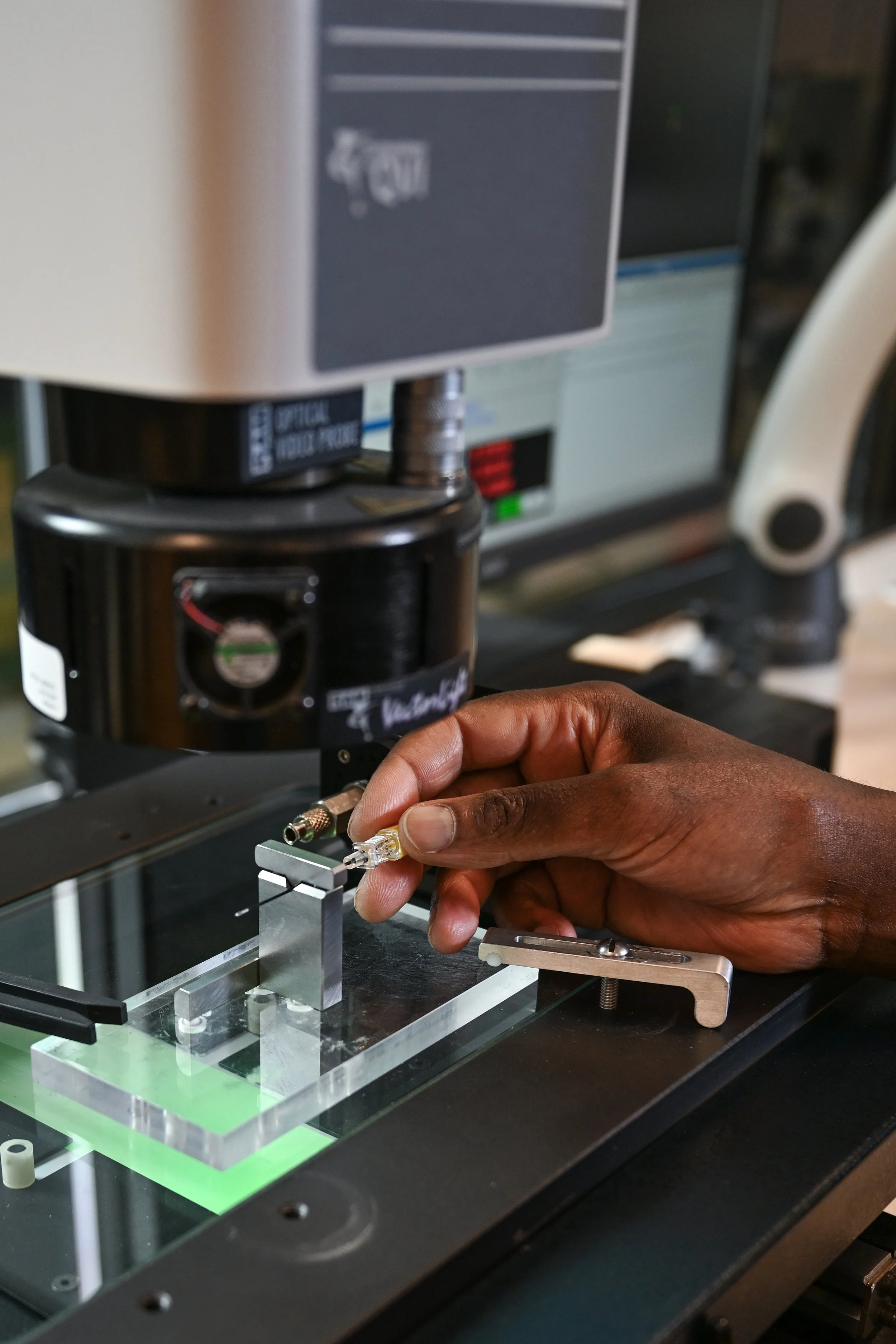 Close-up of a person’s hand positioning a small component under an optical video probe machine for precision inspection in a laboratory or manufacturing setting.