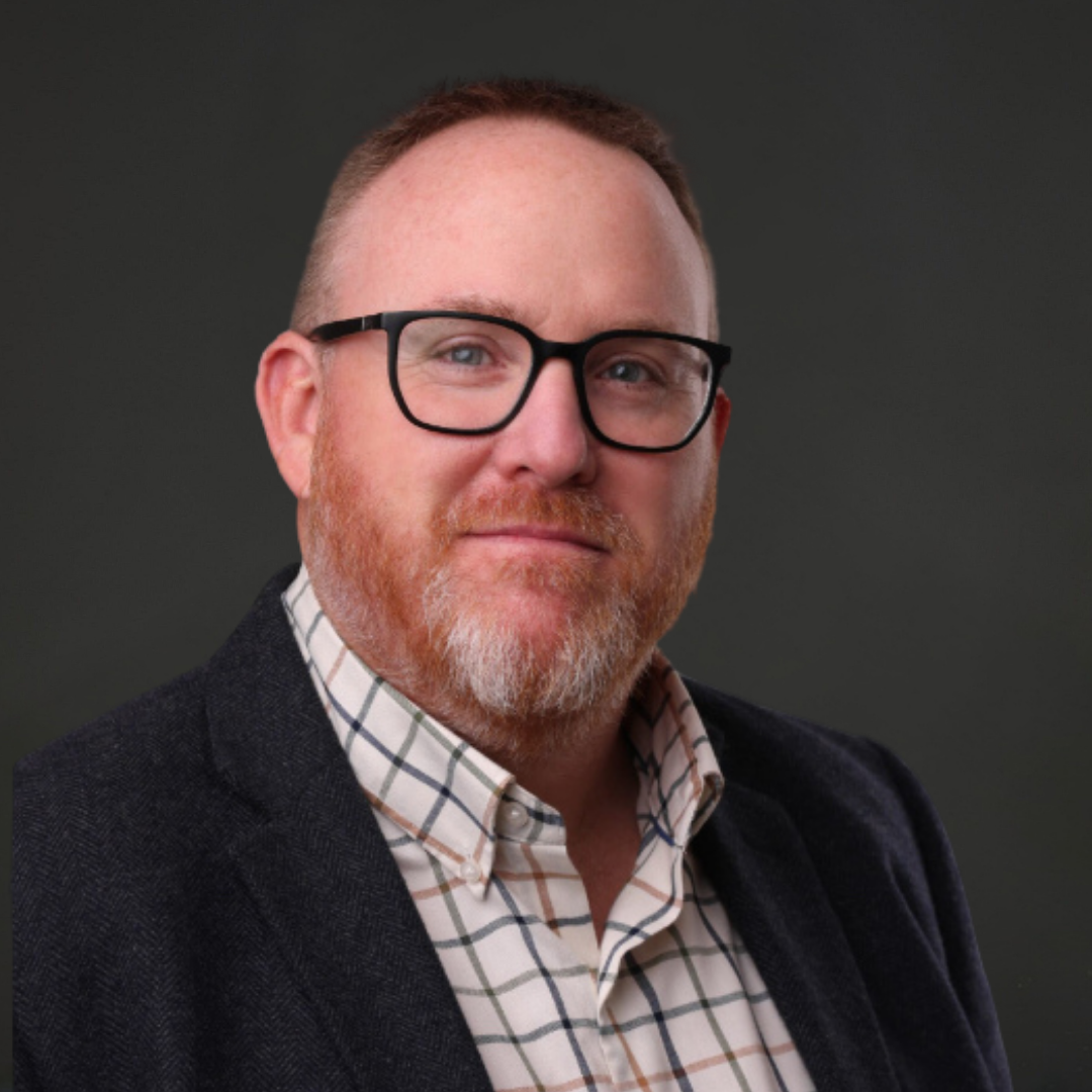Headshot of a man with glasses, red hair, and a beard, wearing a plaid shirt and dark blazer against a dark background.