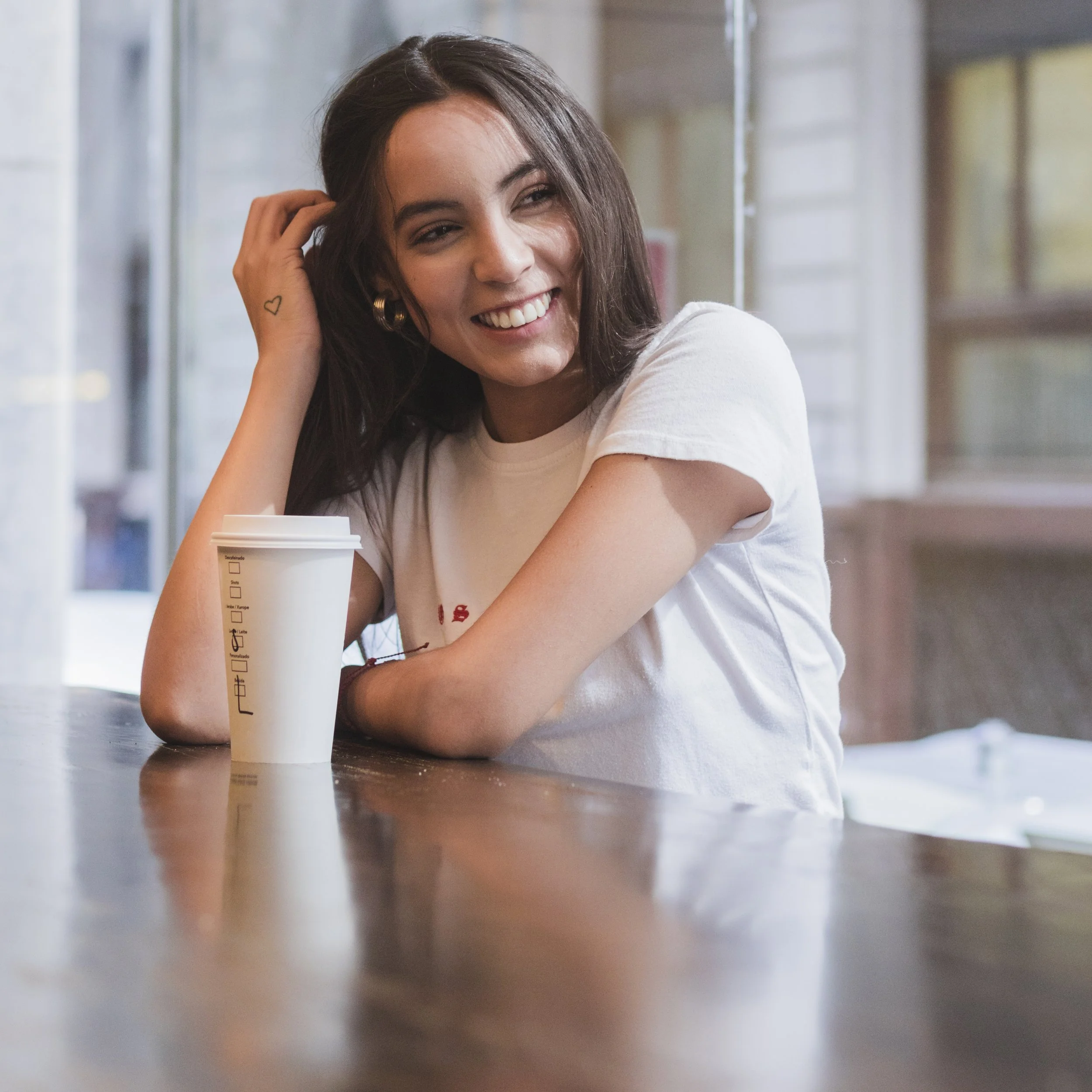 Young woman smiling with a takeaway coffee cup at a wooden table reflecting a moment of ease and connection like those encouraged through Guidance Teletherapy’s corporate wellness webinars