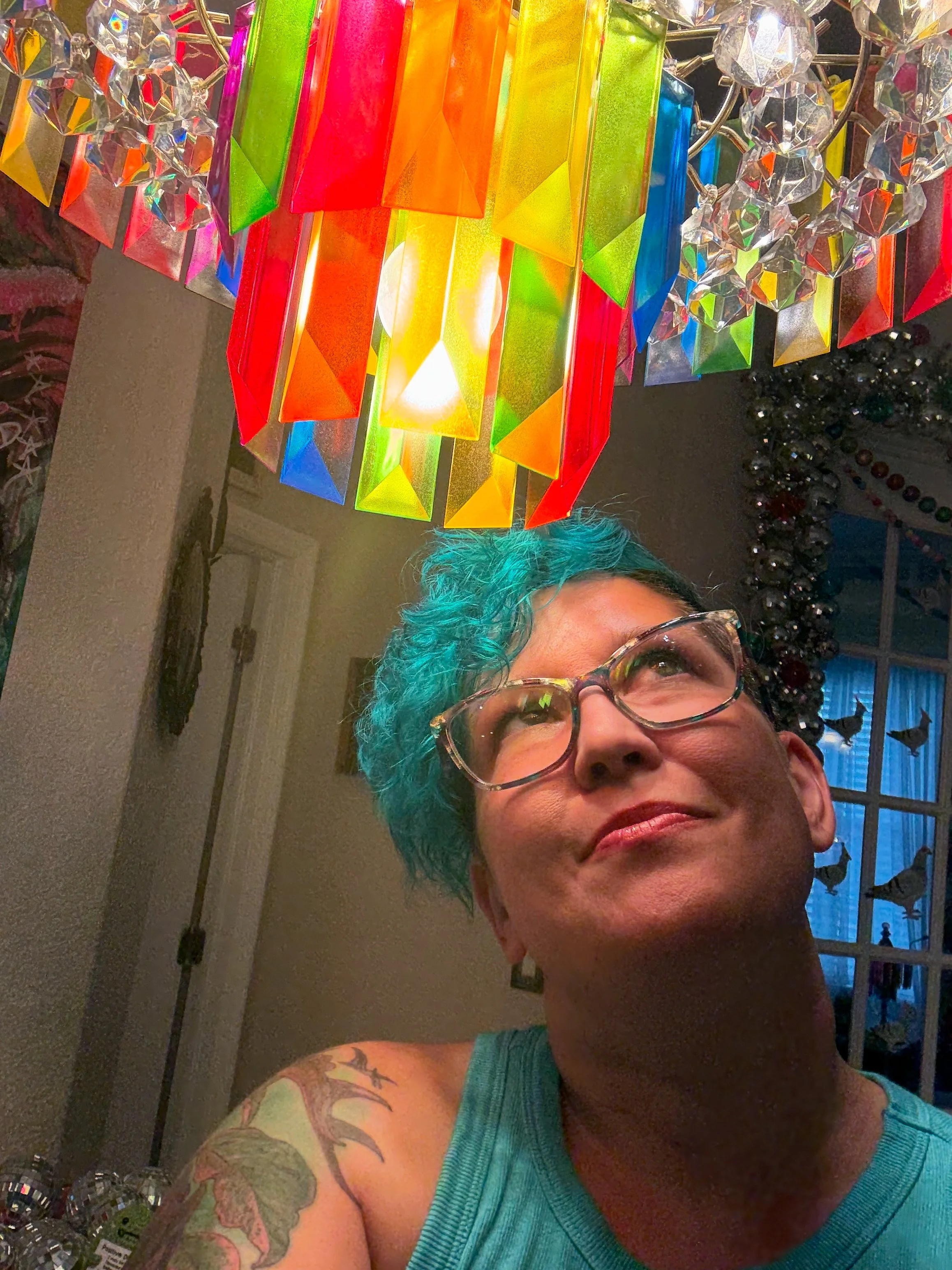 A person with blue hair and glasses looking up at a colorful, multi-colored chandelier hanging from the ceiling.