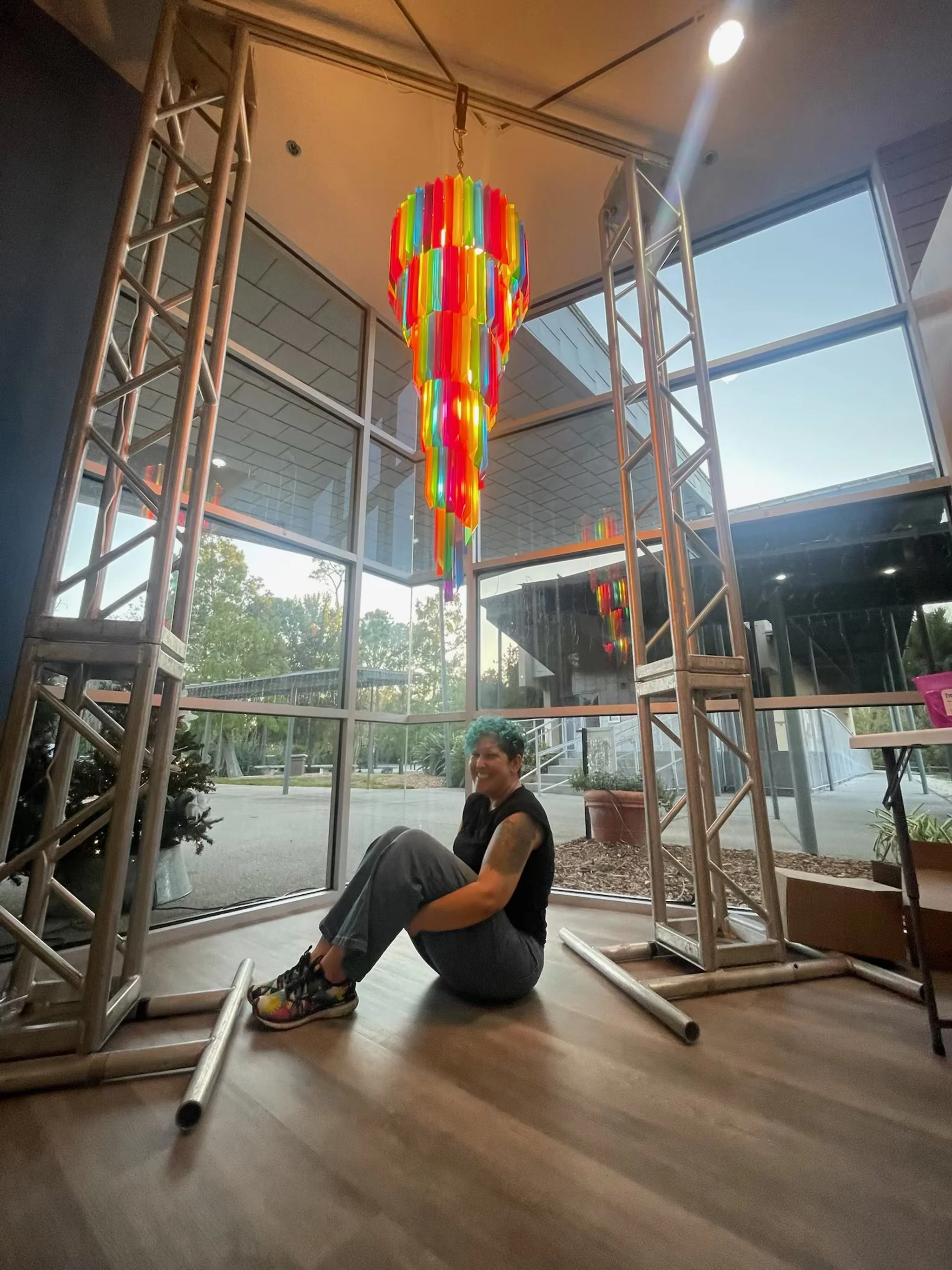 A person sitting on the floor inside a building with large glass windows, a colorful abstract chandelier hanging from the ceiling, and a metal frame structure surrounding them.