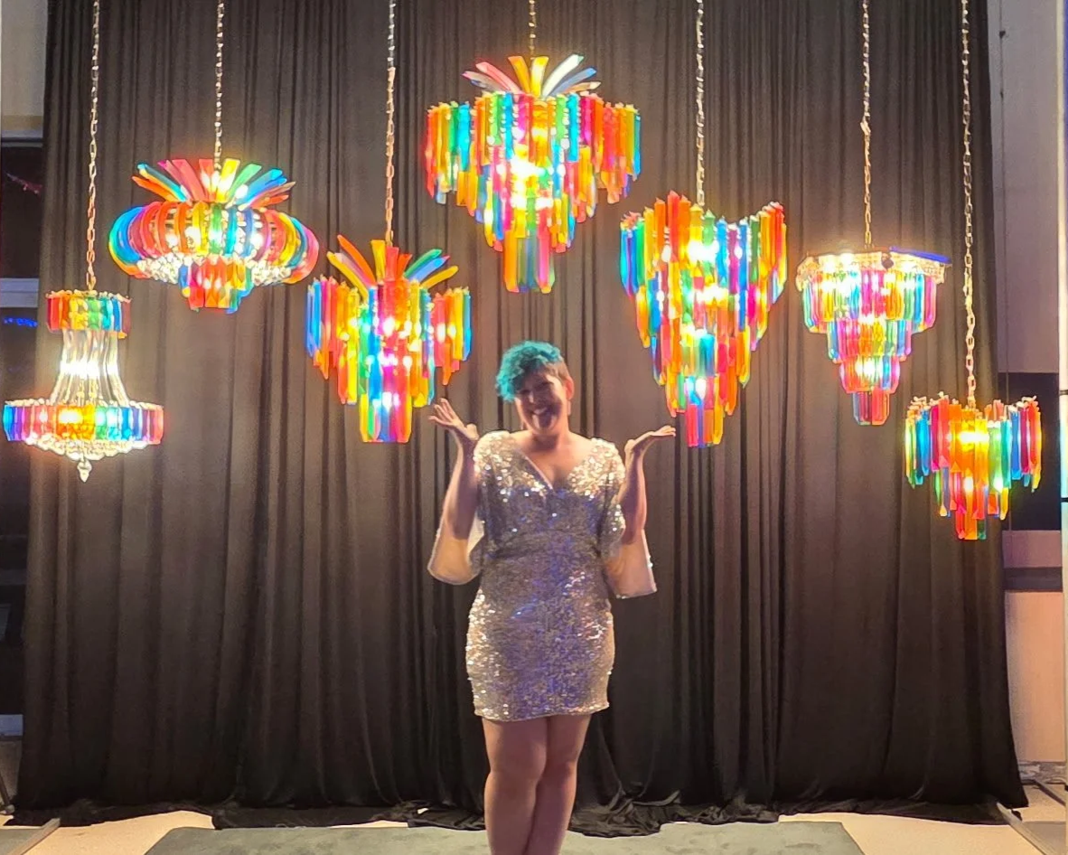 Person with short hair in a shiny, silver dress standing in front of colorful chandelier-style lights hanging from the ceiling, with black curtains in the background.