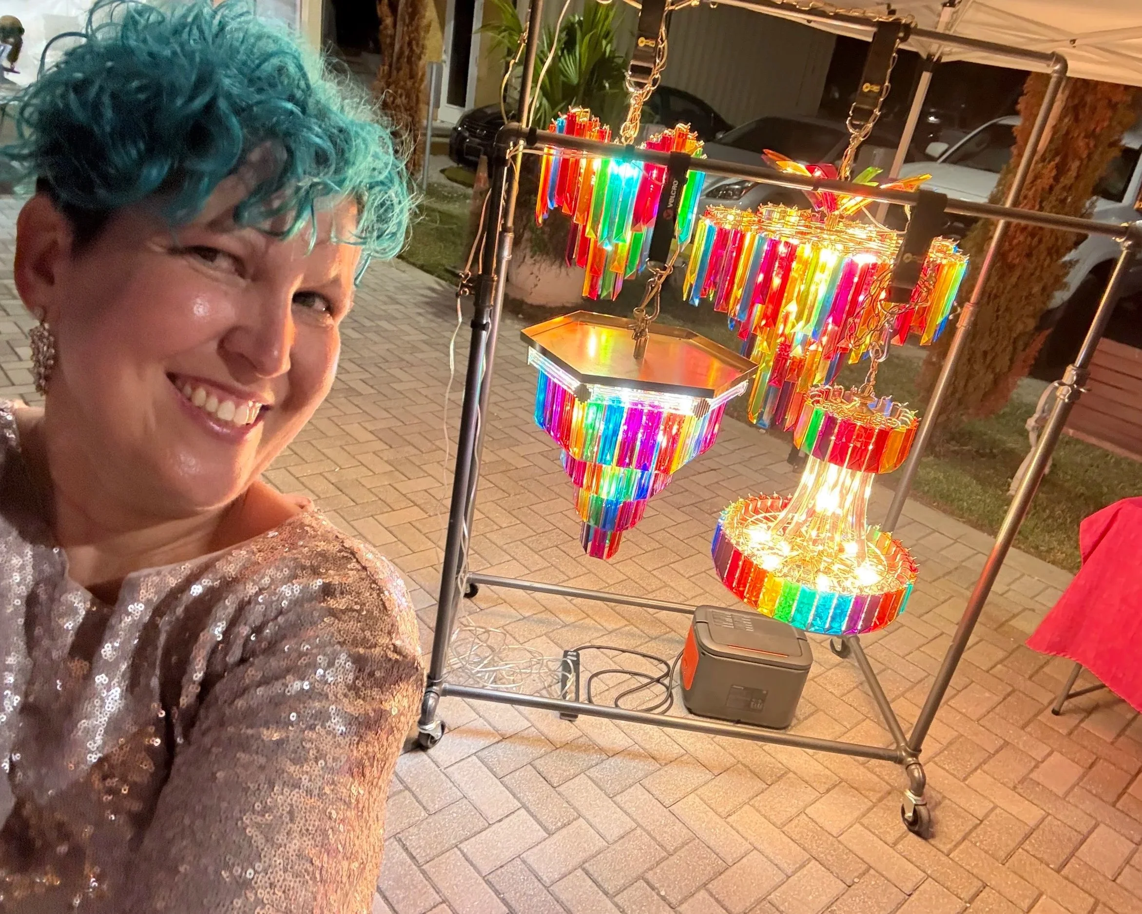 A woman with short teal hair taking a selfie next to a colorful, illuminated art installation made of glow sticks or similar material, displayed on a metal frame outdoors at night.