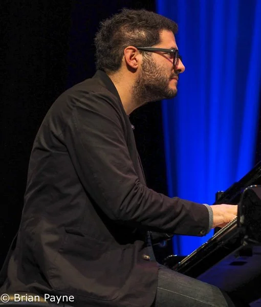 A man playing a grand piano on stage, wearing glasses and a dark jacket, with a blue curtain backdrop.