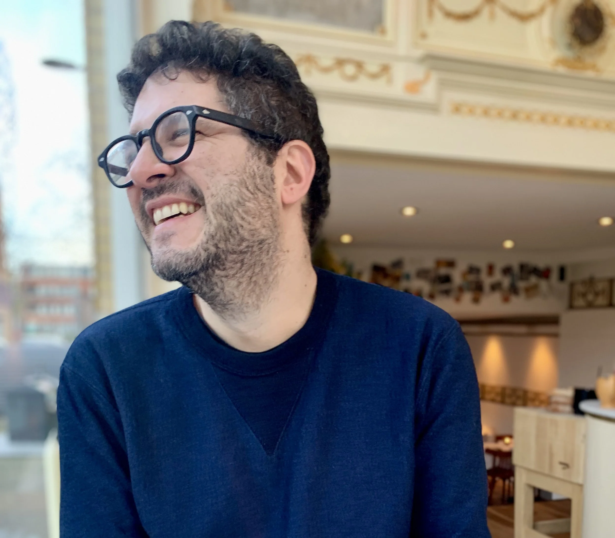 A person with glasses smiling in an indoor setting with beige walls and framed pictures.
