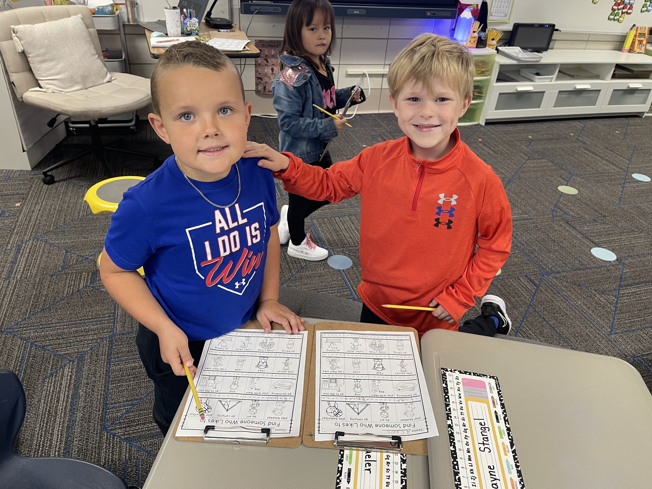 Two young boys are standing at a table in a classroom, smiling at the camera, with worksheets in front of them. A girl in the background holds a pencil and looks away. The classroom has educational materials and supplies around.