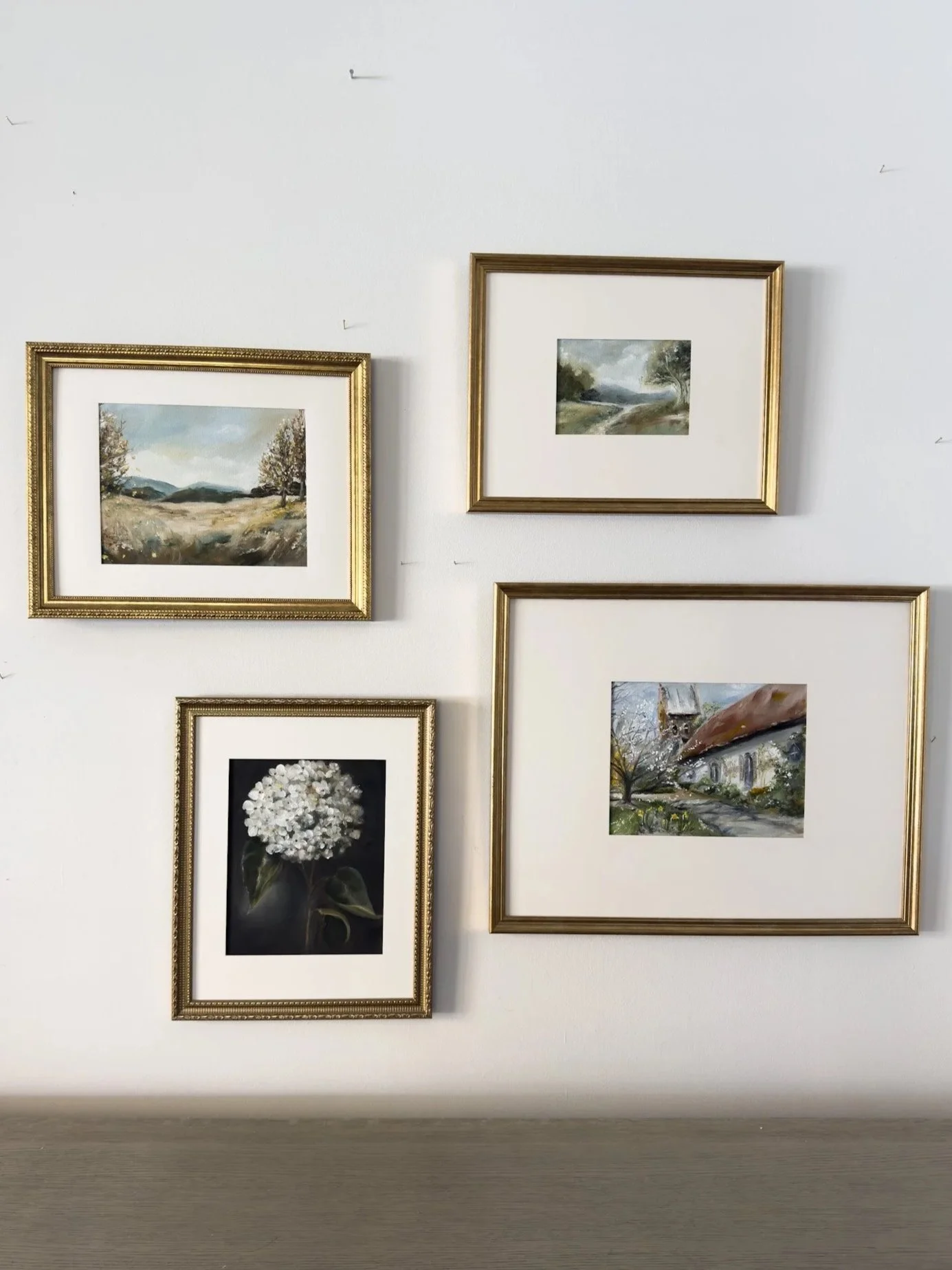 A white wall with four framed pictures, three landscape paintings and one photograph of a white flower, arranged in a cluster.