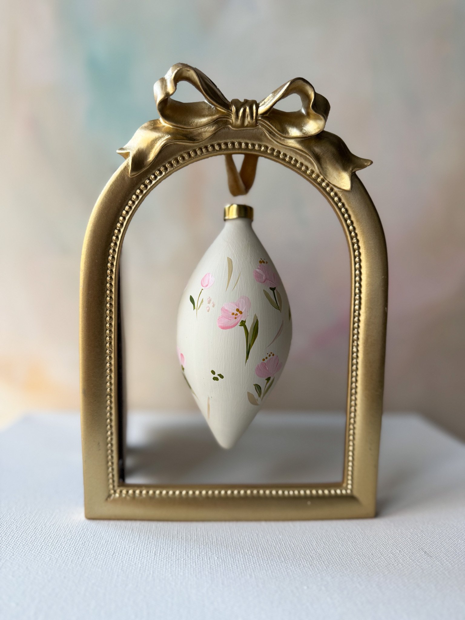 Decorative hanging ornament with floral design inside a gold frame with a bow on top.