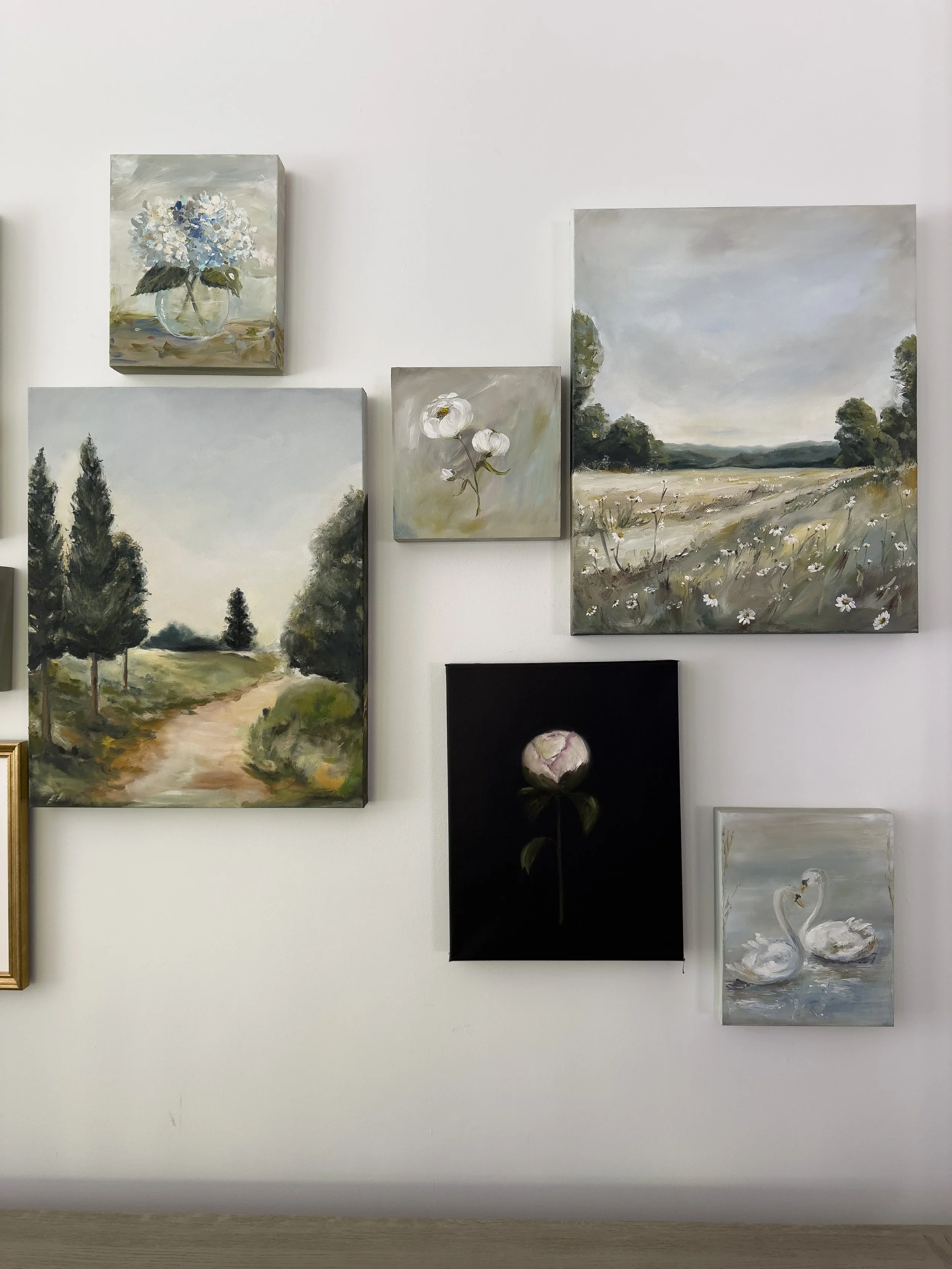 Collection of landscape and floral paintings hung on a white wall, including images of trees, fields, flowers, and swans.