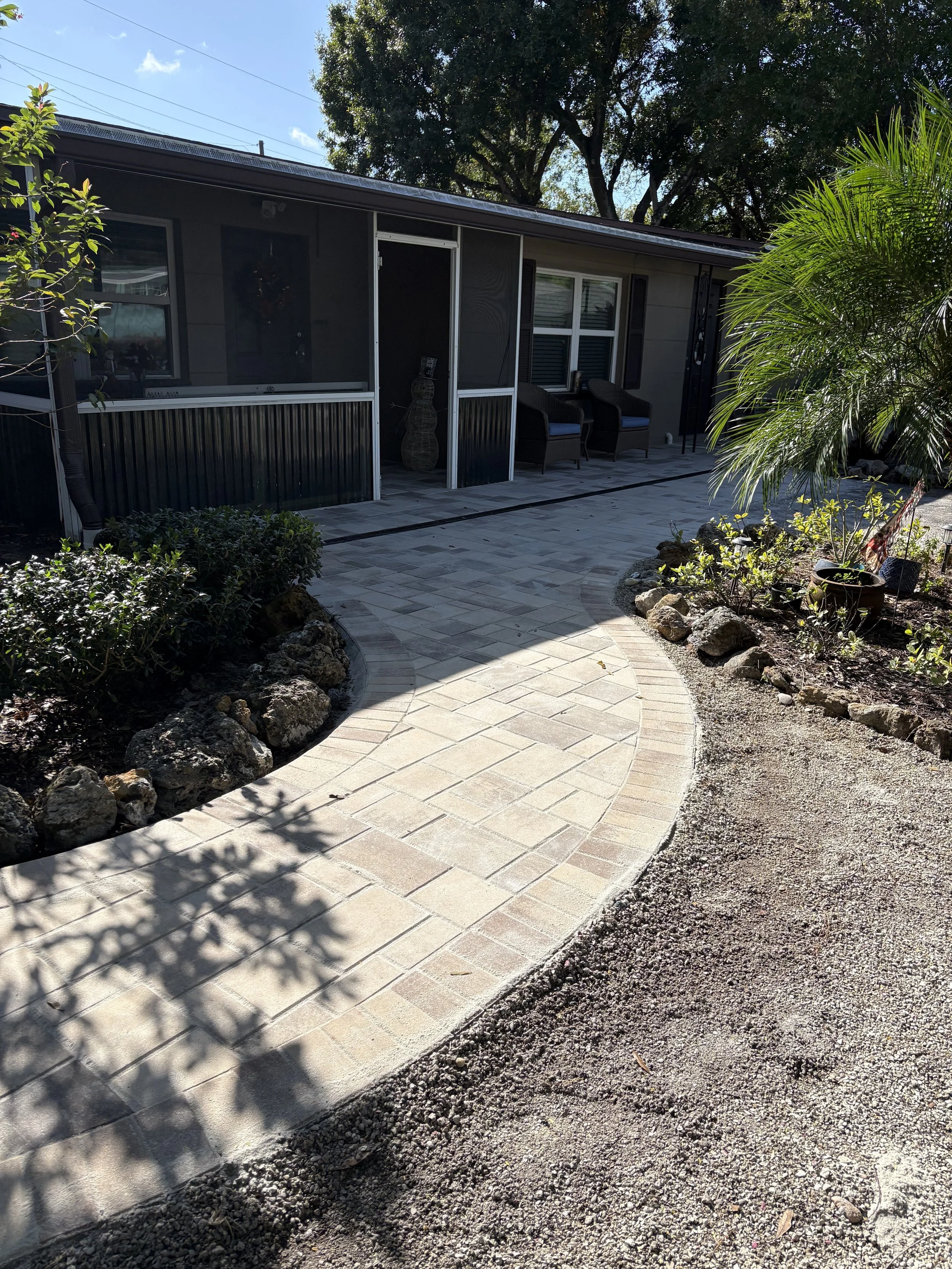 A backyard patio with stone pavers, surrounded by landscaped garden beds with rocks and bushes. There are outdoor chairs and a screened porch attached to the house, with trees and blue sky in the background.