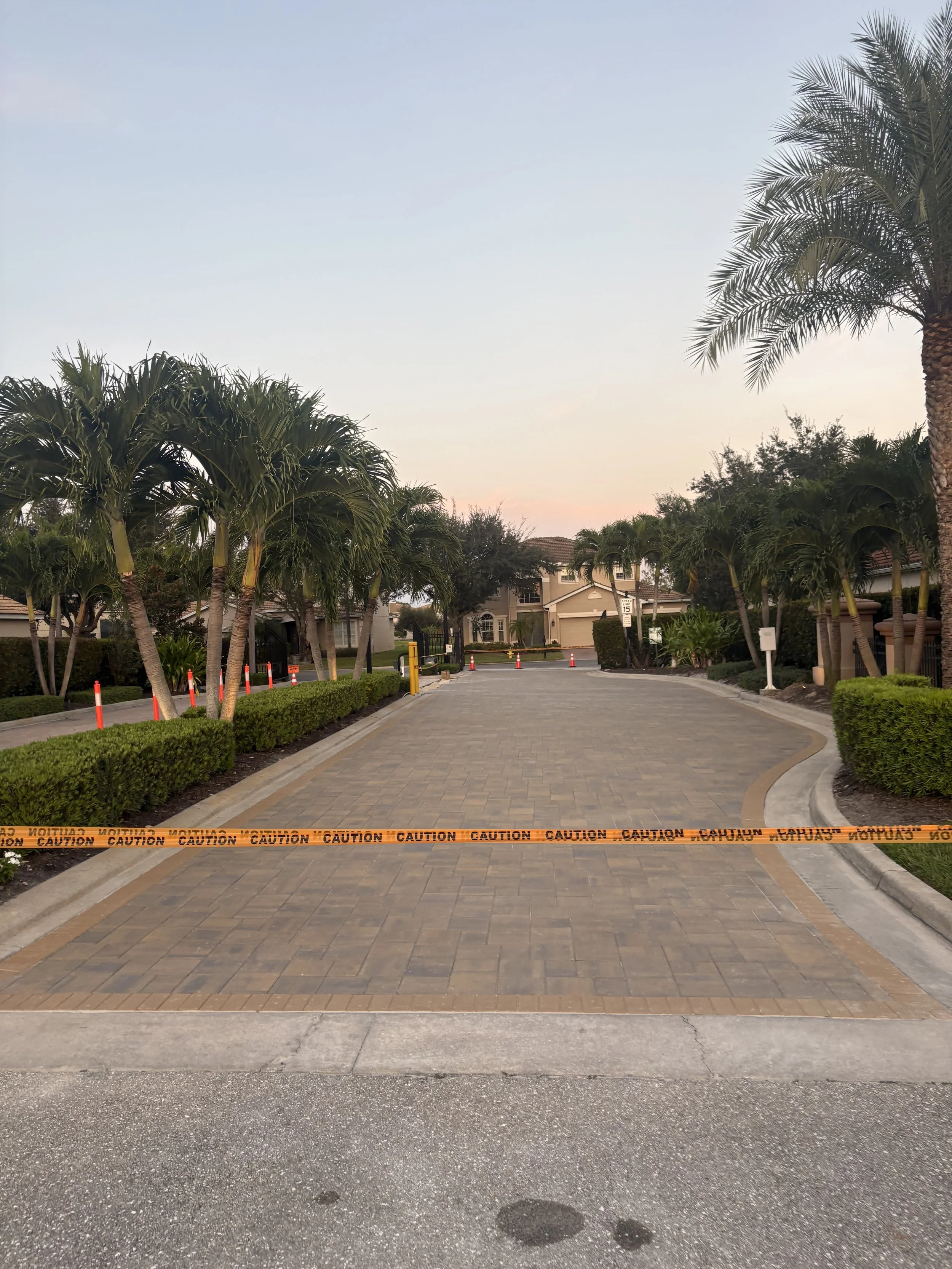 Residential street blocked by caution tape with palm trees and houses visible in the background.