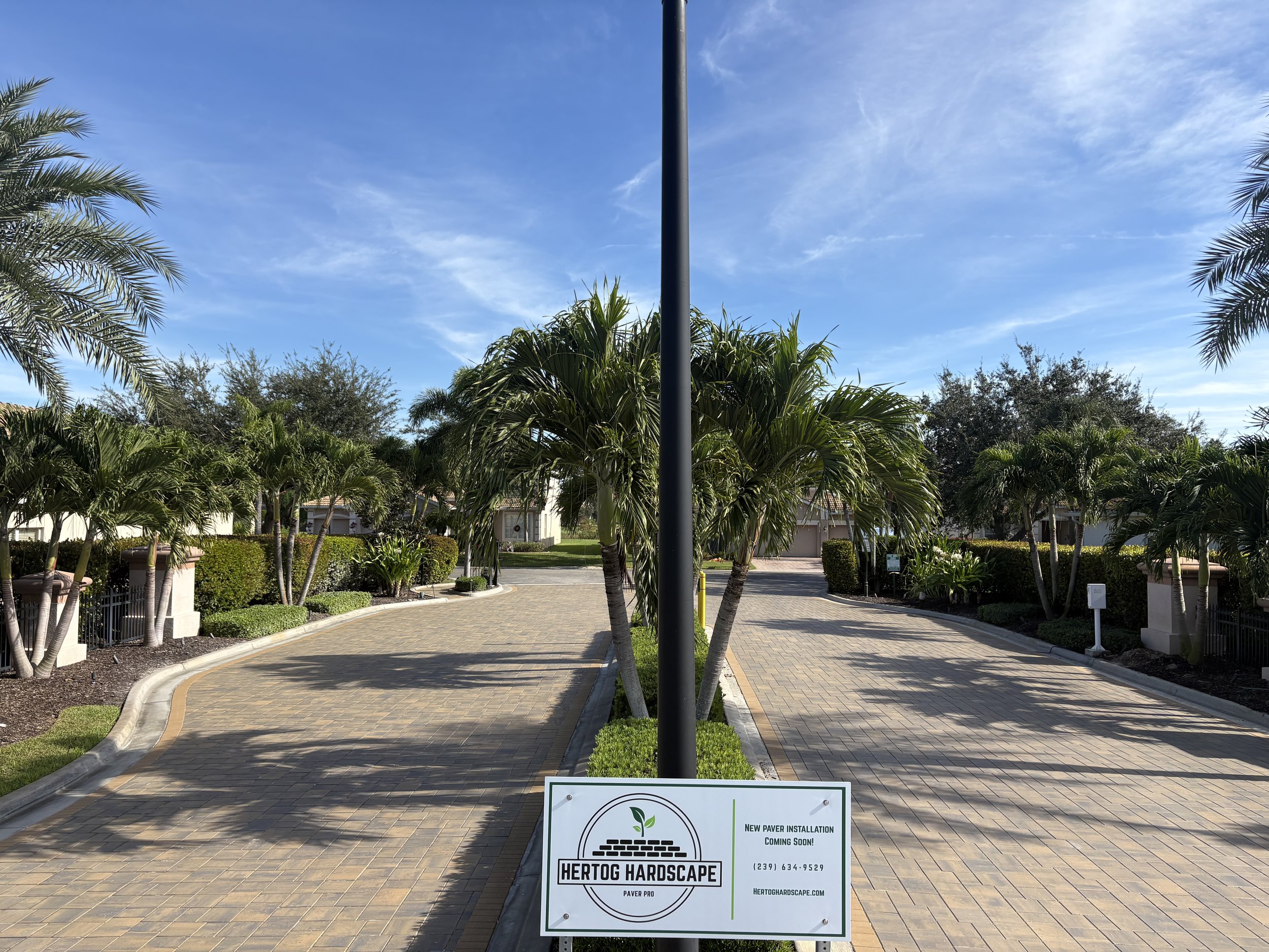 Residential street with paved walkway, palm trees, and landscaped bushes under a blue sky with some clouds. A sign in the foreground reads 'Hertog Hardscape' and mentions upcoming paver installation.