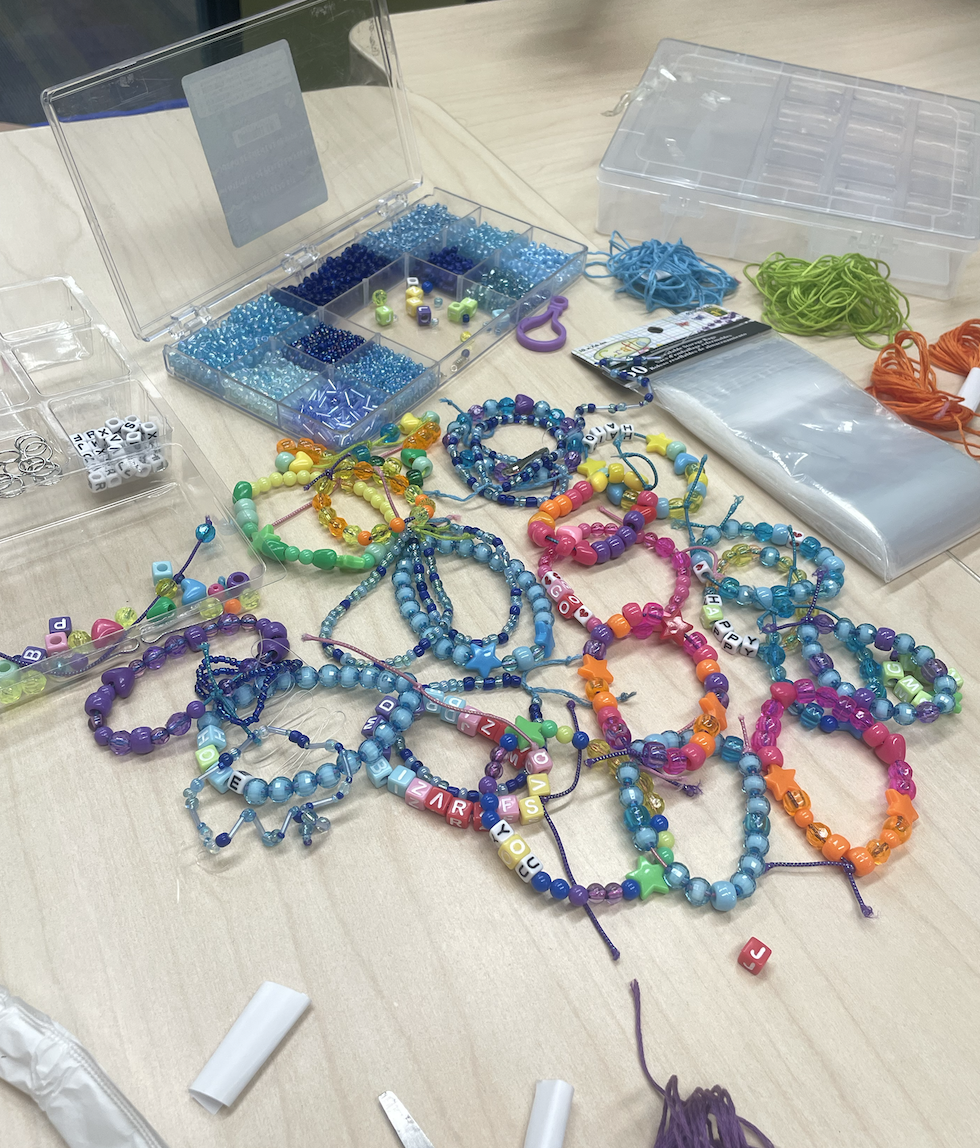 Colorful bead necklaces and bracelets made with various beads, some with alphabet letters, are displayed on a table along with beading supplies such as thread, scissors, and storage containers.