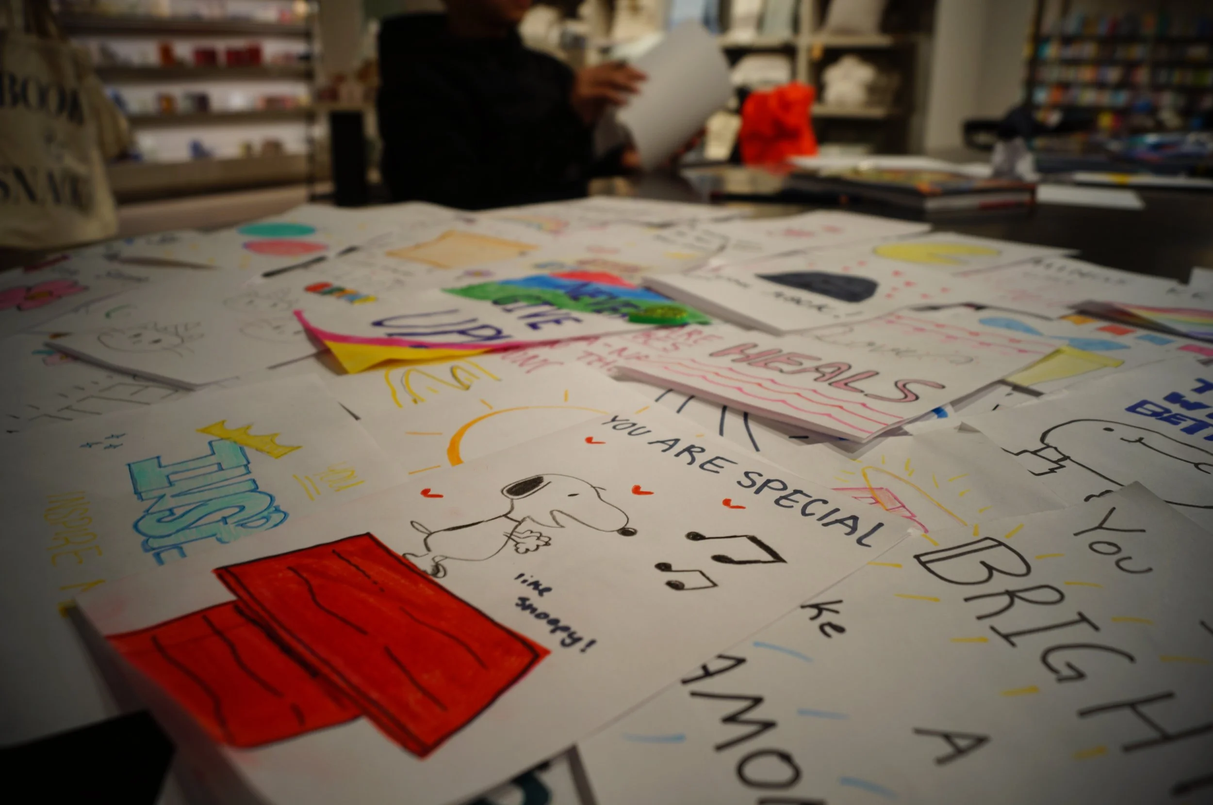 A table covered with colorful handmade cards and drawings, with a person in the background reading a book or paper in a bookstore or library.