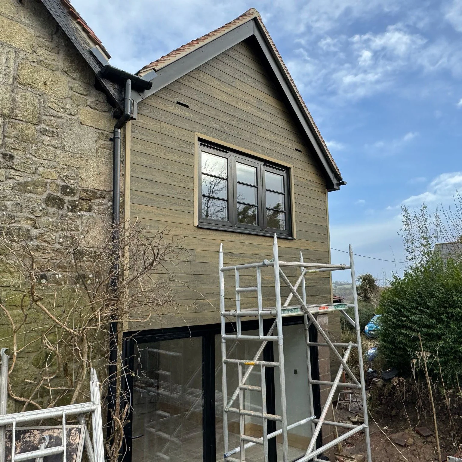 House extension during construction with wood siding and scaffolding, adjacent to stone building.