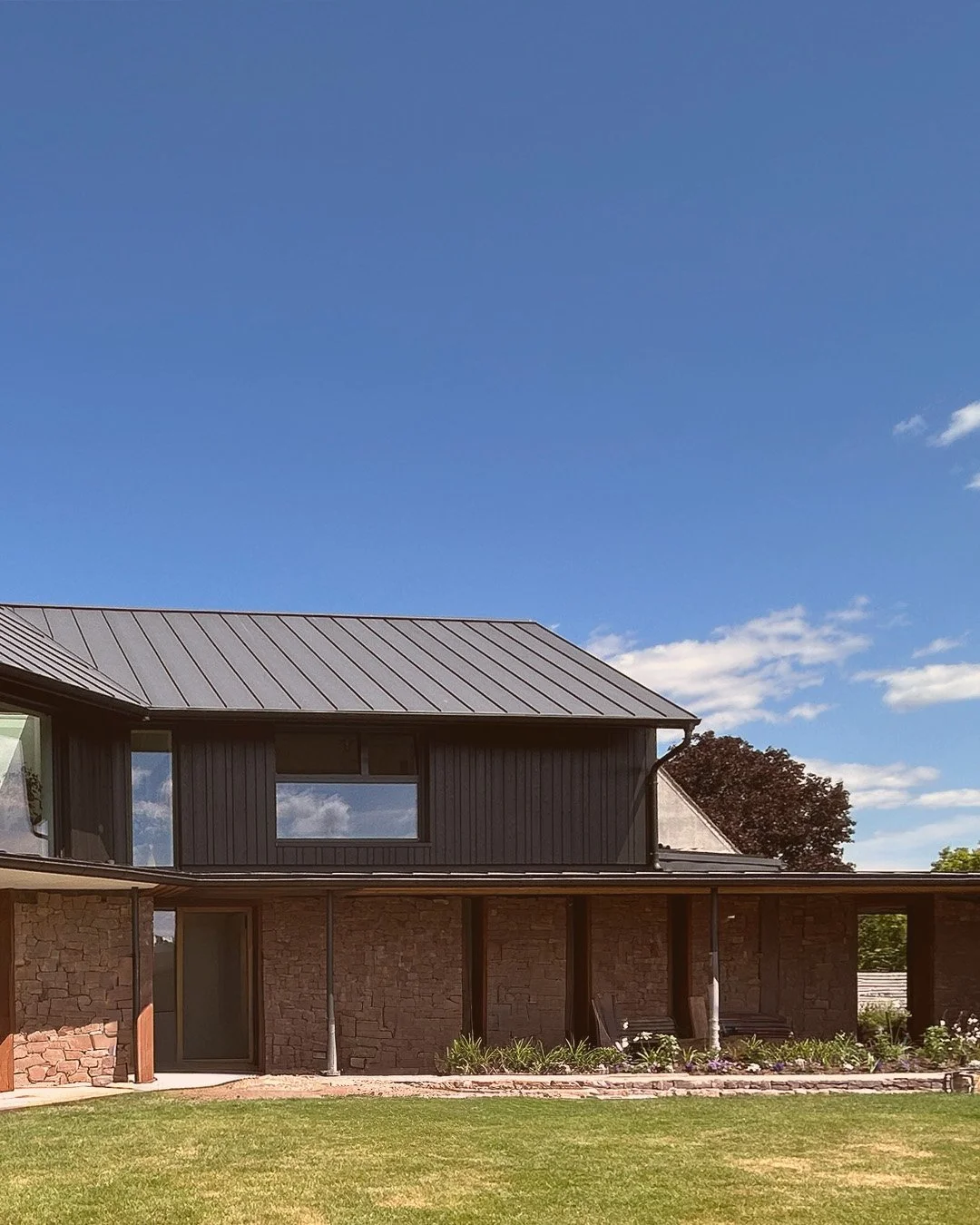 Folia Architects - Modern house with brick and dark panel exterior, metal roof, and landscaped yard under a clear blue sky.