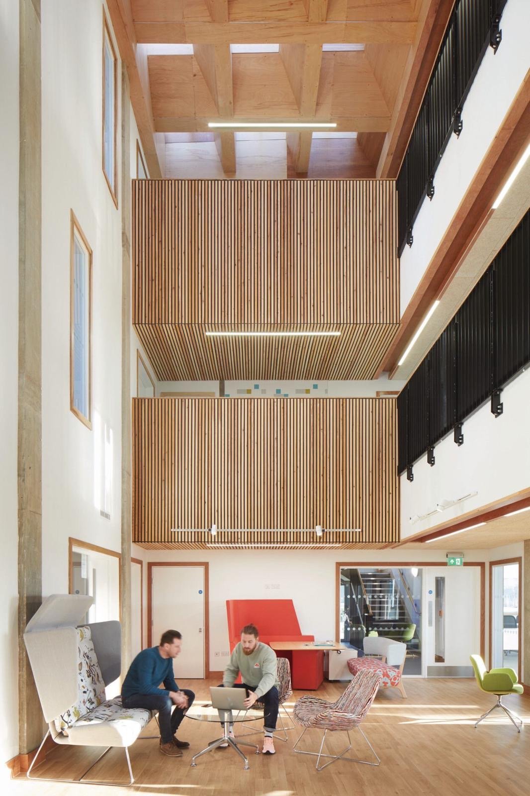 A spacious indoor area with high ceilings, wooden beams, and modern furniture. Two men are sitting at a small round table with a laptop, engaged in conversation. The room features colorful chairs, a red booth seat, and large windows allowing natural light.