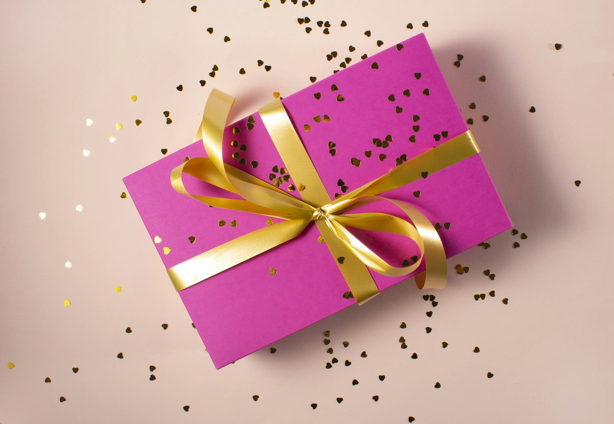 Pink gift box with a gold ribbon and bow, decorated with small black heart-shaped confetti, on a light pink surface.