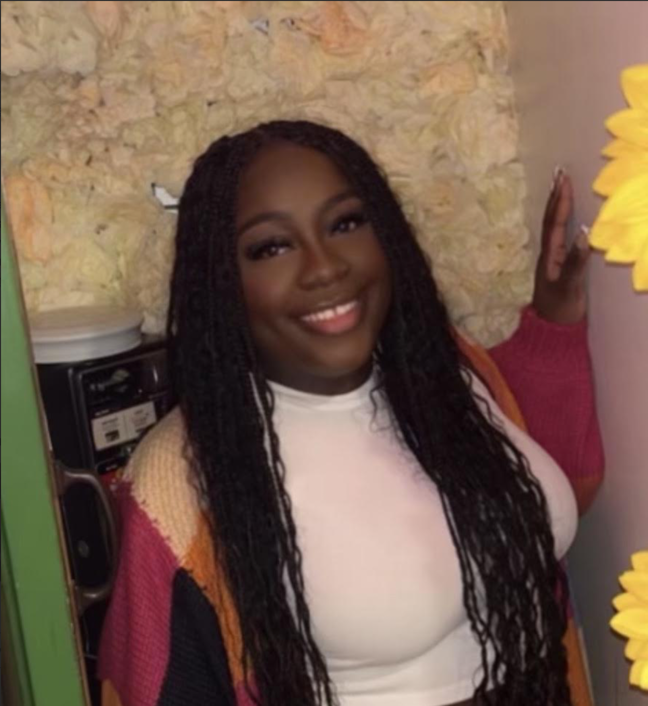 A smiling woman with long dark braided hair wearing a white shirt and a colorful cardigan, standing inside a room with a stone wall and a yellow sunflower decoration.