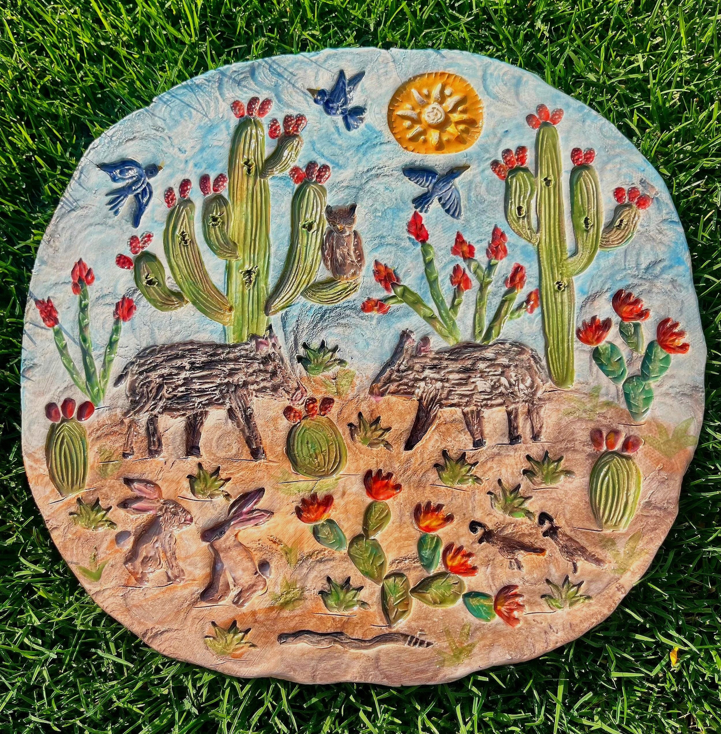 A decorative round ceramic plaque featuring a desert scene with cacti, javelinas, birds, owls, rabbits, and scorpions, surrounded by colorful flowers. The sun is also depicted in the sky. The plaque is placed on green grass.