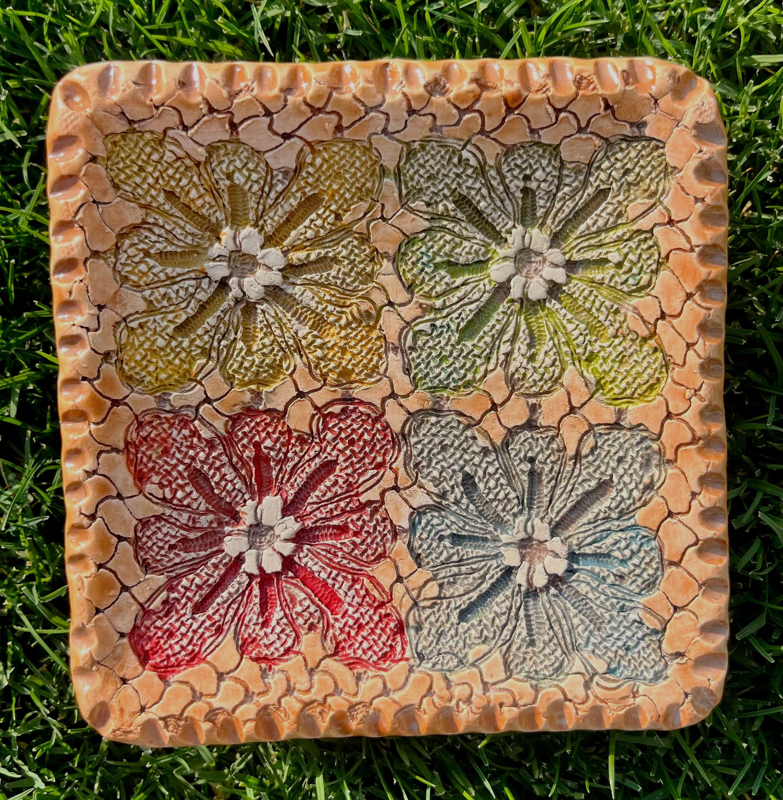 Handmade ceramic dish with four textured floral patterns in green, red, yellow, and blue on a cracked stone-like surface, displayed on grass.