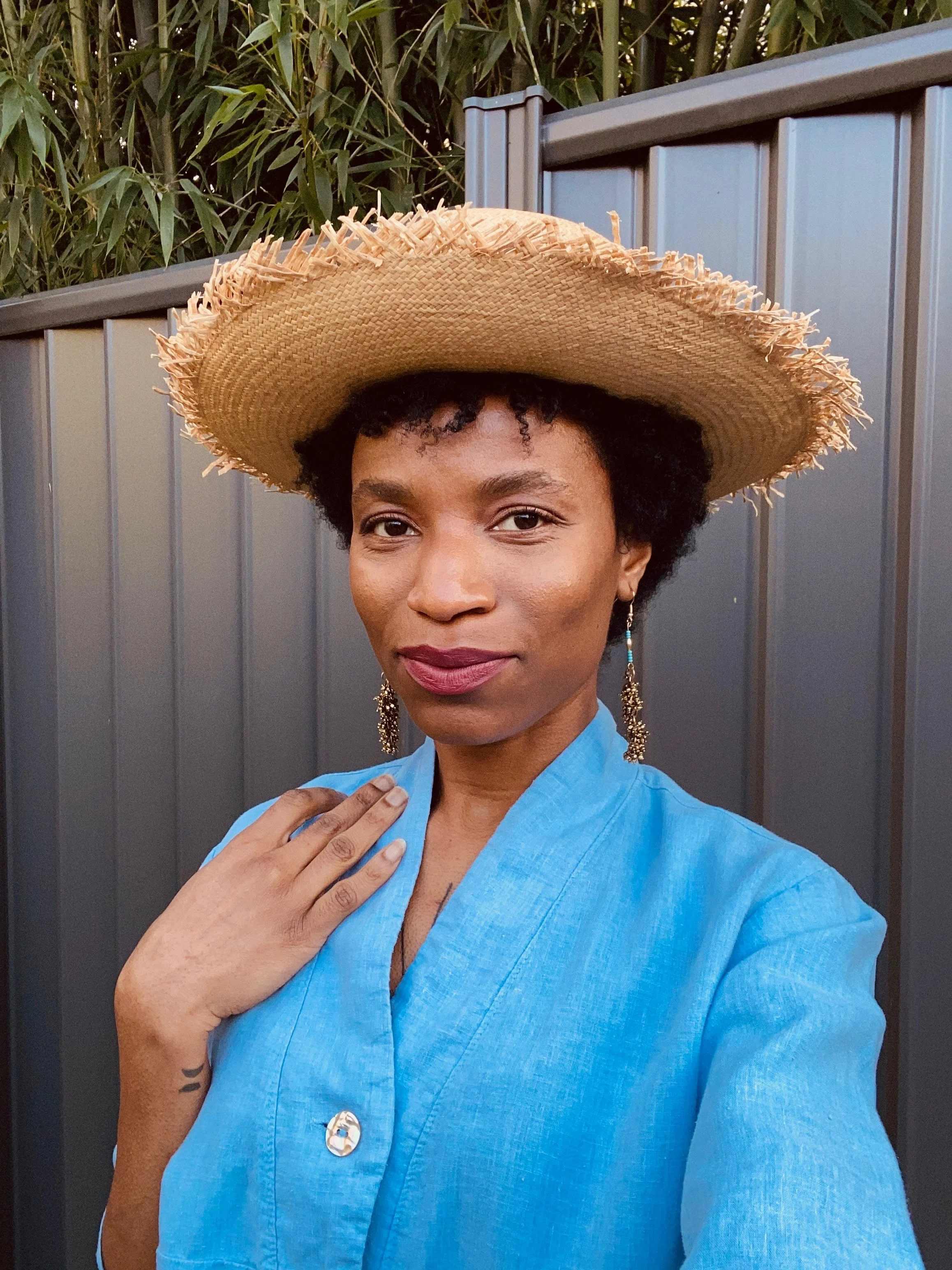 Person wearing a blue shirt and straw hat, standing in front of a metal fence with greenery in the background.