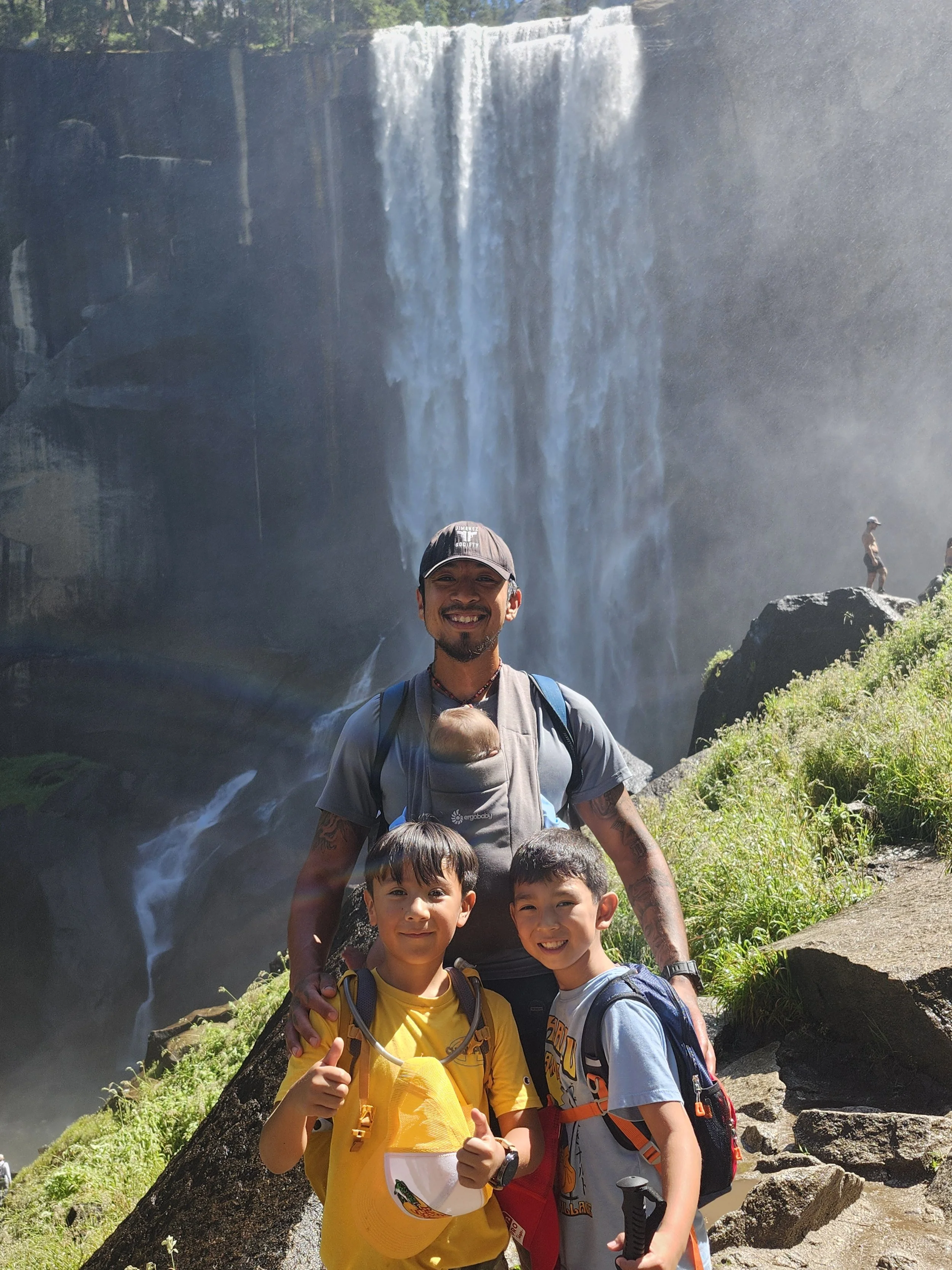 A man and three children smiling at the camera in front of a waterfall at a scenic outdoor location, with rocks and greenery around.