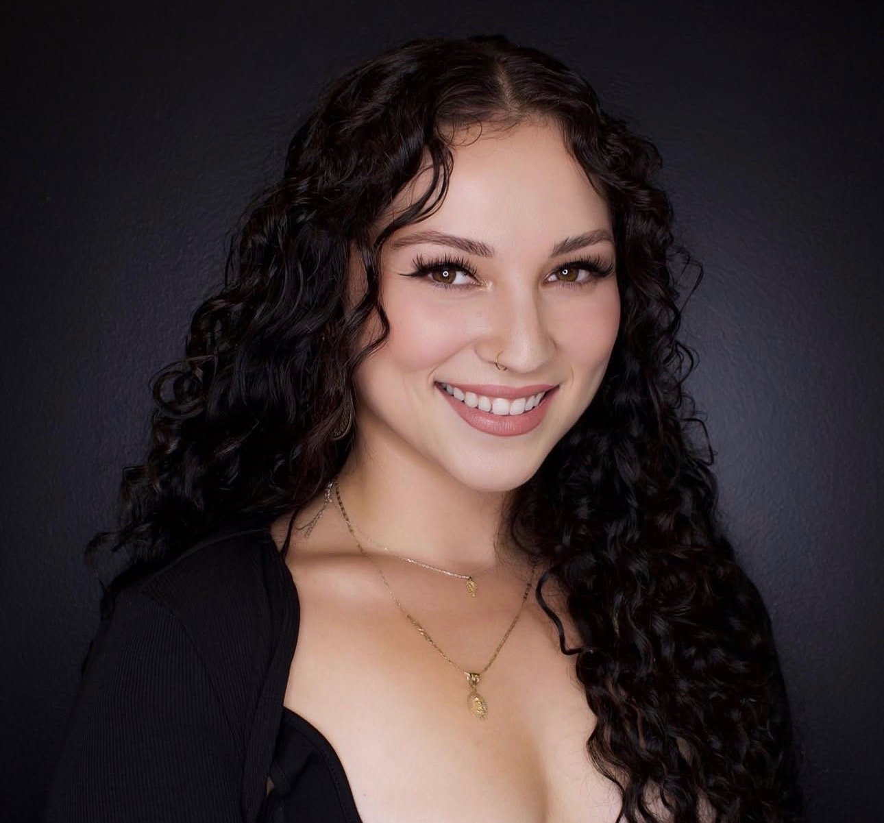 A young woman with long, curly black hair smiling at the camera, wearing a black top and layered gold necklaces, against a dark gray background.
