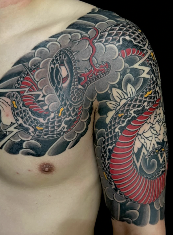 Tattoo of a Japanese-style dragon on a person's shoulder and upper chest, featuring intricate patterns, black and red colors, clouds, and floral elements.