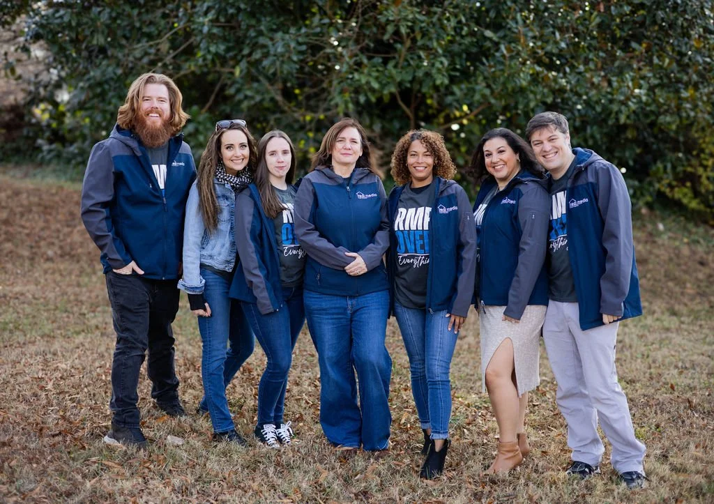 Group of seven people standing outdoors on grass with trees in the background, wearing matching navy blue jackets and posing for a photo, smiling at the camera.