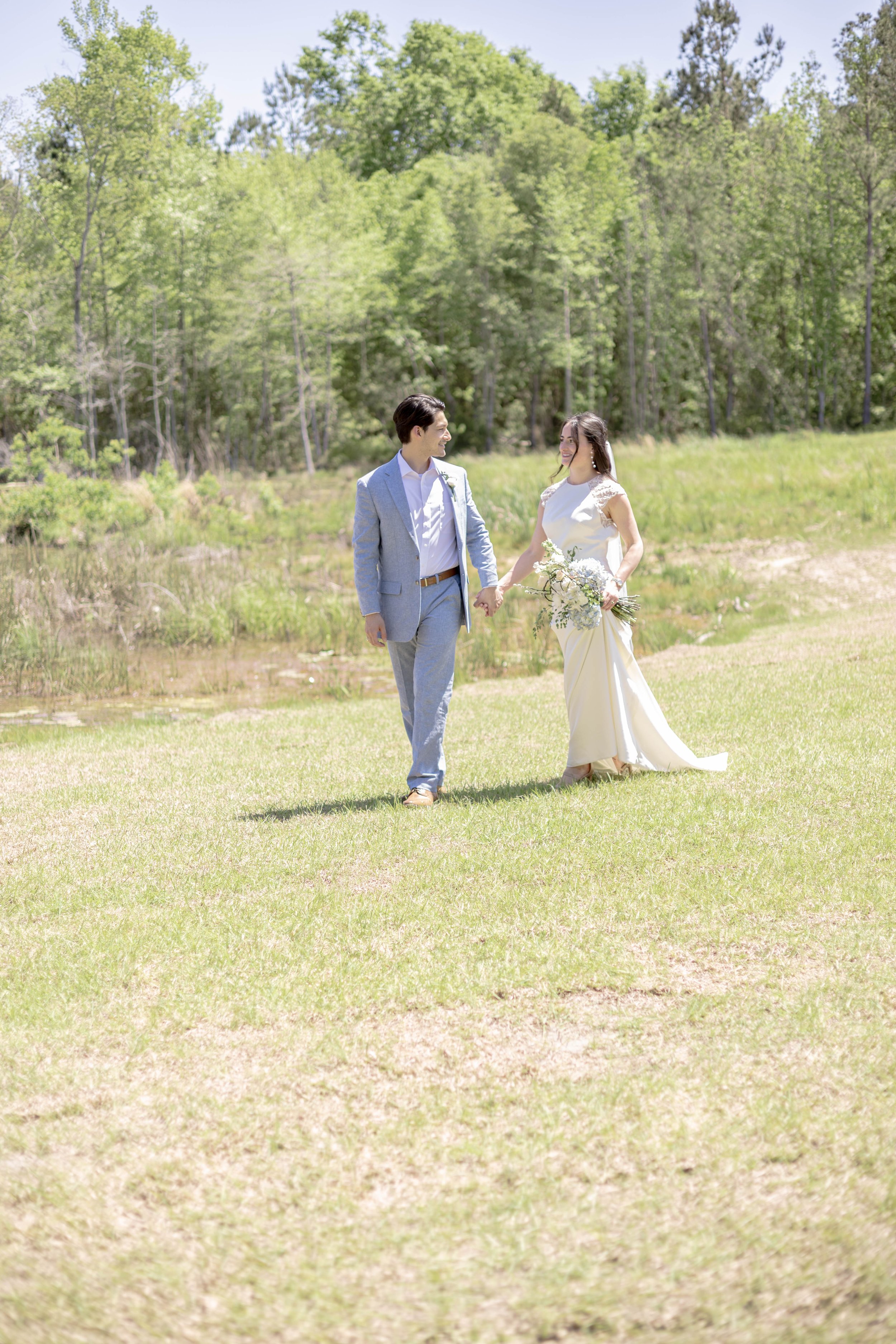 A couple walking hand in hand on a grassy field, the woman wearing a white dress and holding a bouquet, the man in a light gray suit, with trees in the background.