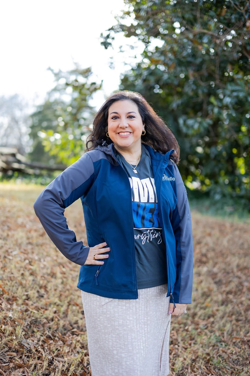 A woman with dark hair, wearing a blue jacket over a graphic t-shirt, standing outdoors in a grassy area with trees in the background. She is smiling and has one hand on her hip.