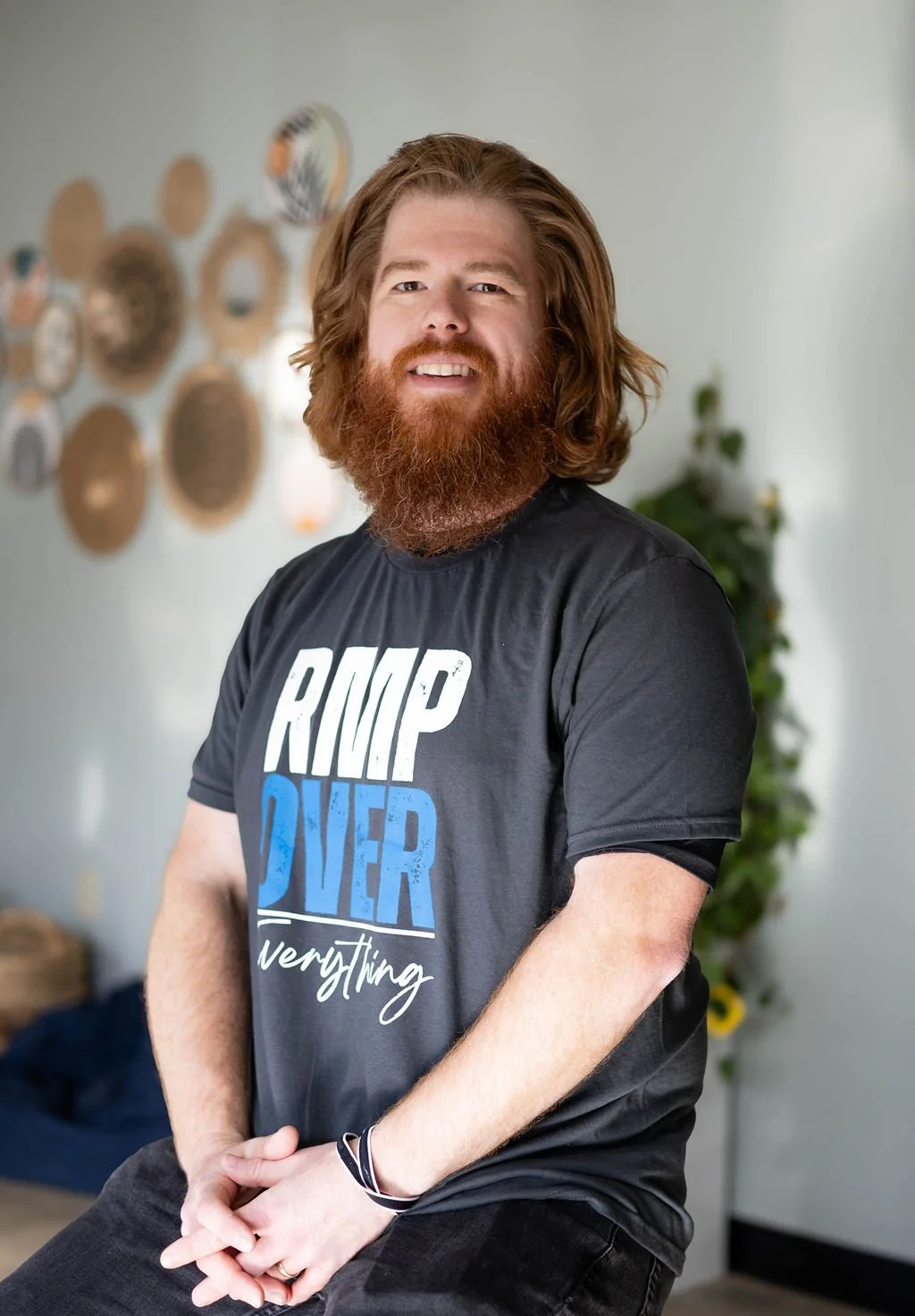 A man with long red hair and a beard smiling, wearing a black t-shirt, standing indoors with wall art in the background and a potted plant.