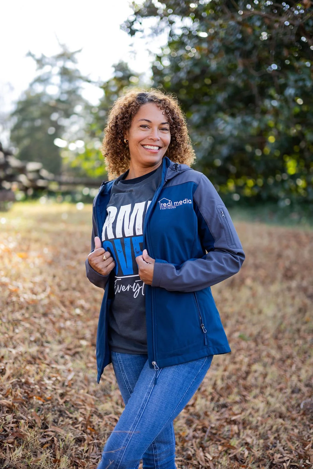 A woman smiling outdoors in a park during fall, wearing a blue jacket and a T-shirt, with trees and fallen leaves in the background.