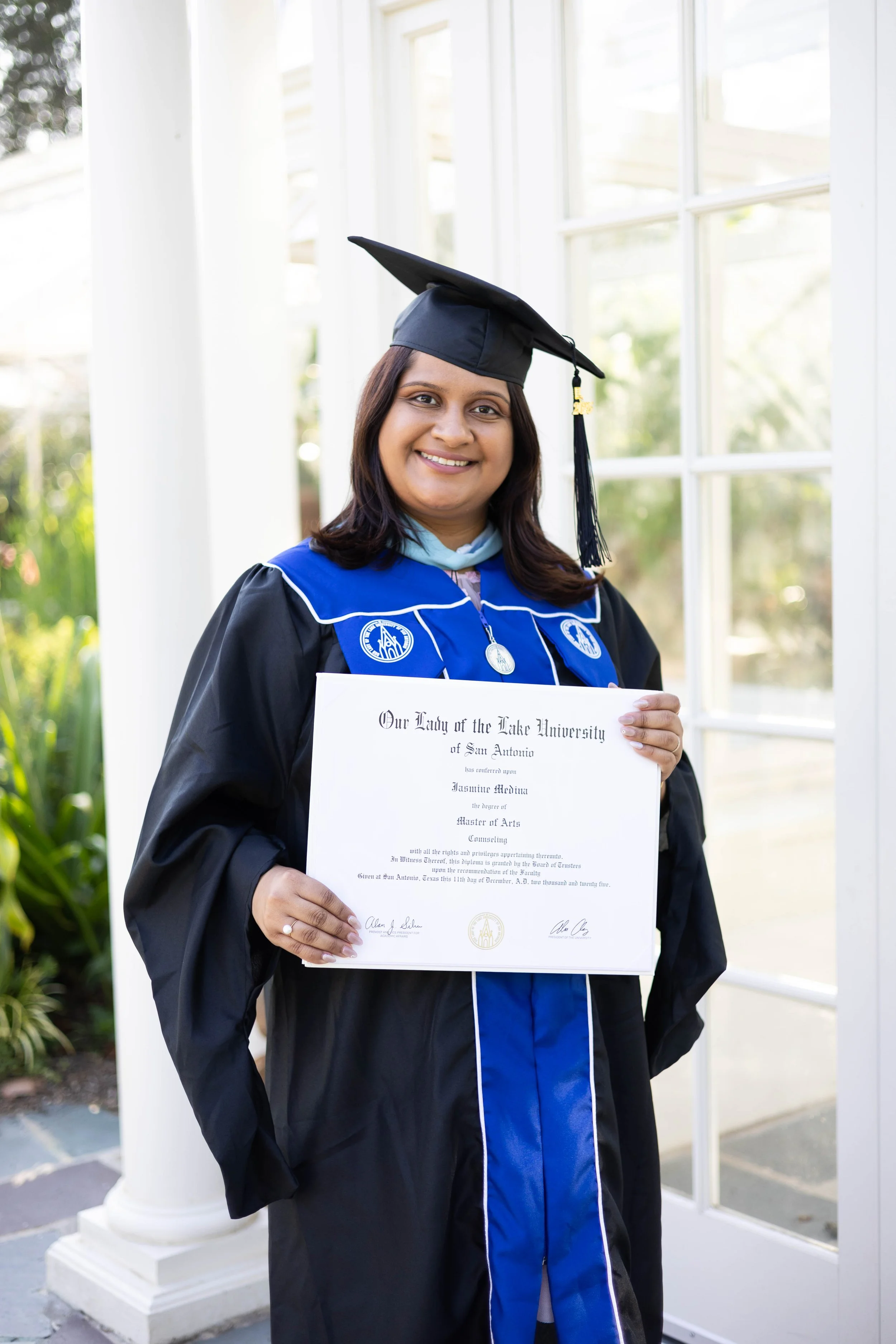 Smiling woman in graduation gown and cap holding a diploma in front of a white column and windows, outdoors in daylight.