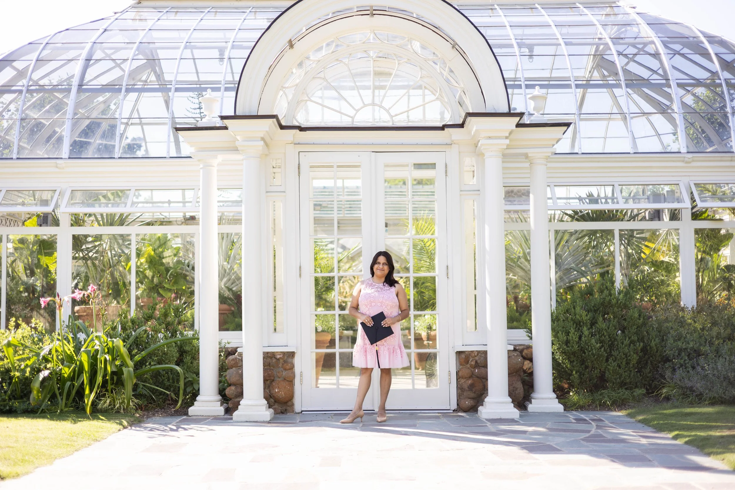 Woman in a pink floral dress holding a black folder, standing in front of a white greenhouse with glass panels and columns, surrounded by greenery and plants.