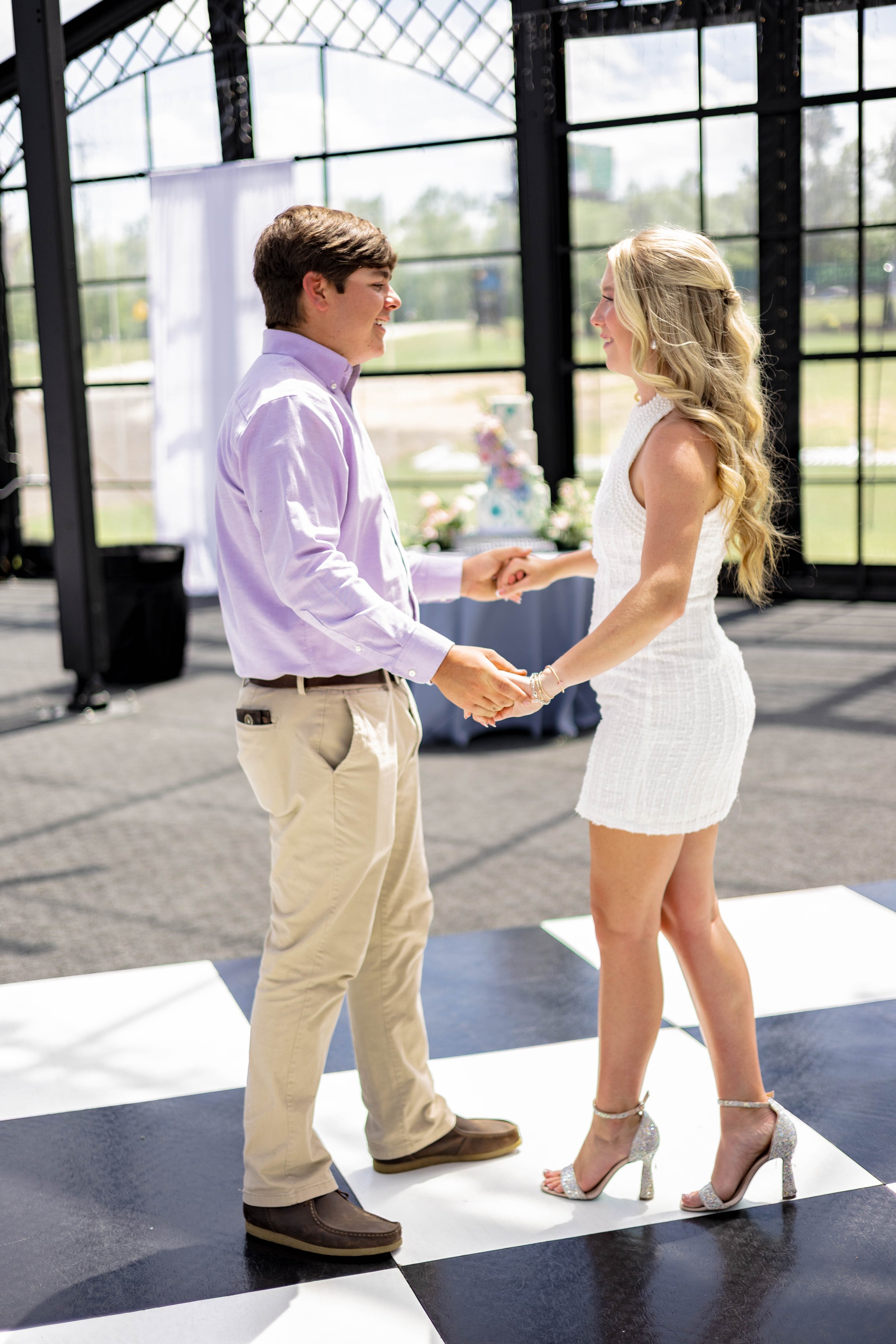 A man in a purple shirt and khaki pants dances with a woman in a white dress and silver high heels on a black and white checkered floor, inside a glass-walled venue.